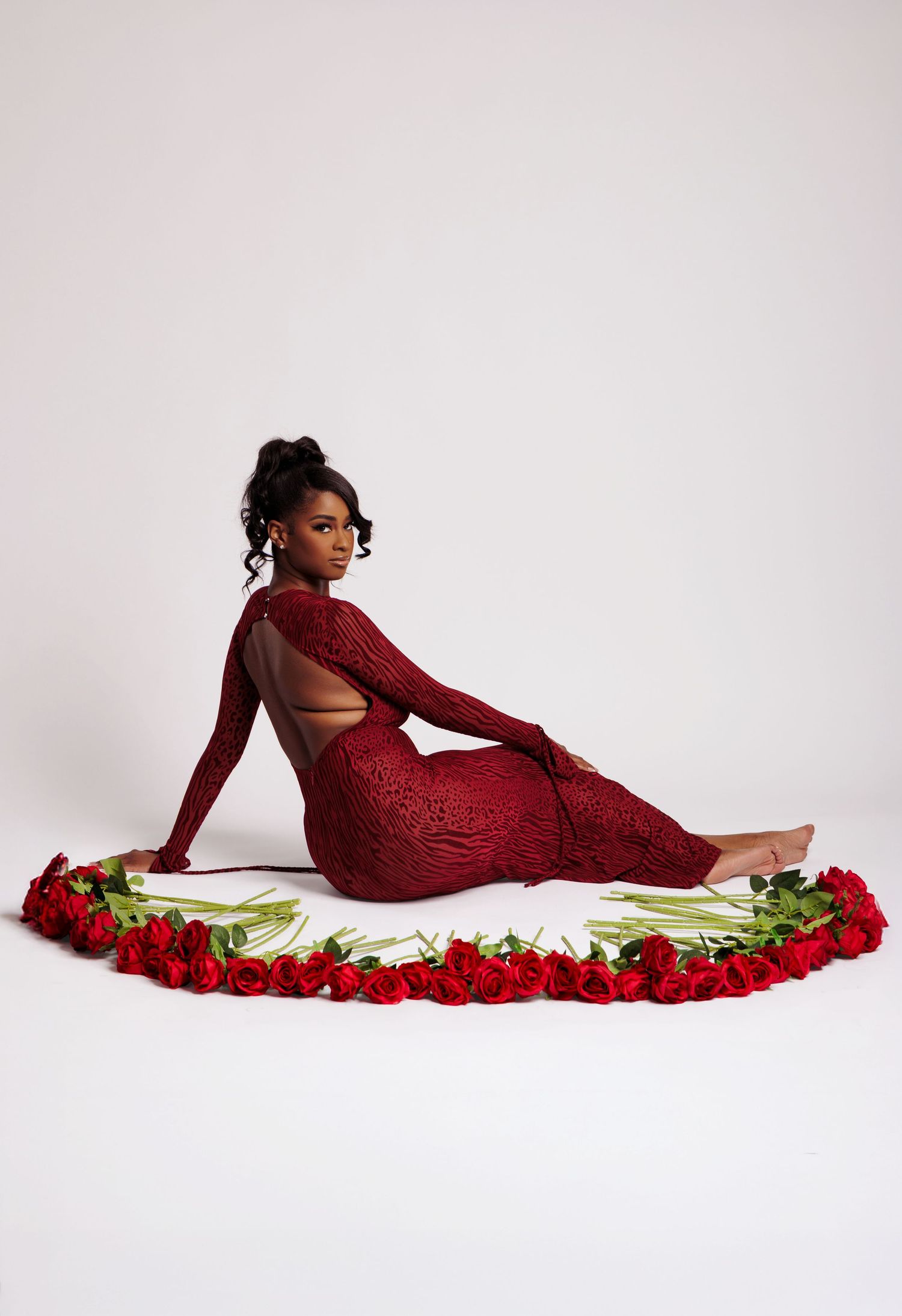 Woman in red lace gown poses elegantly surrounded by red roses on white background.