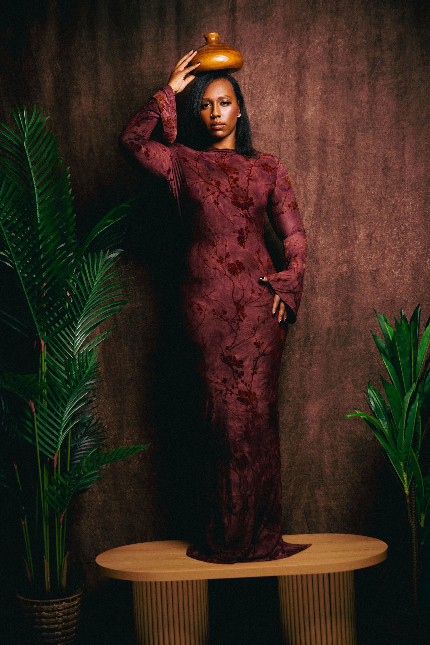 Woman in burgundy lace gown balancing gold vase on head, standing on wooden platform with tropical plants.