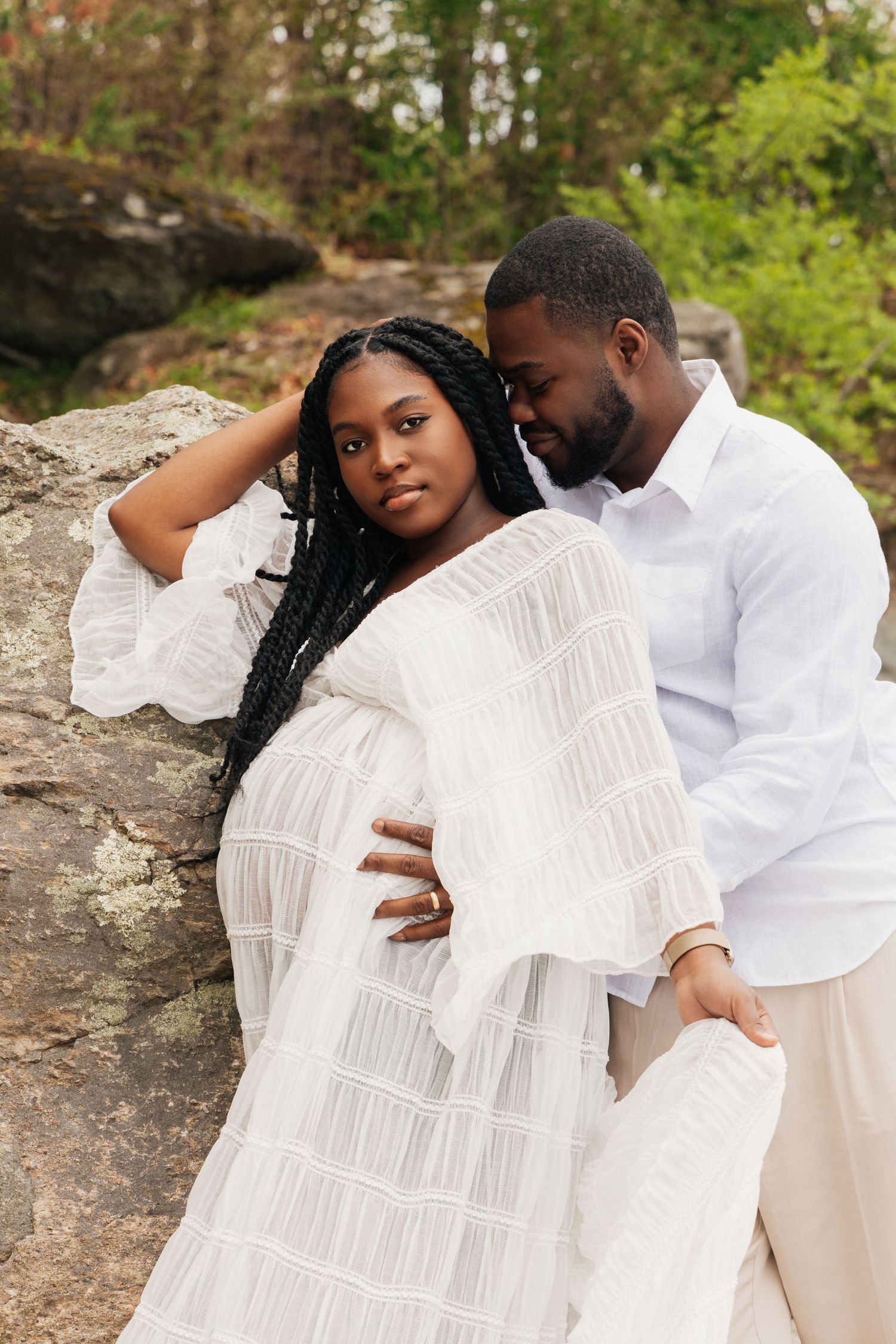 Expecting Black couple leaning against large rock outdoors, man in white shirt tenderly embracing pregnant woman in white dress.