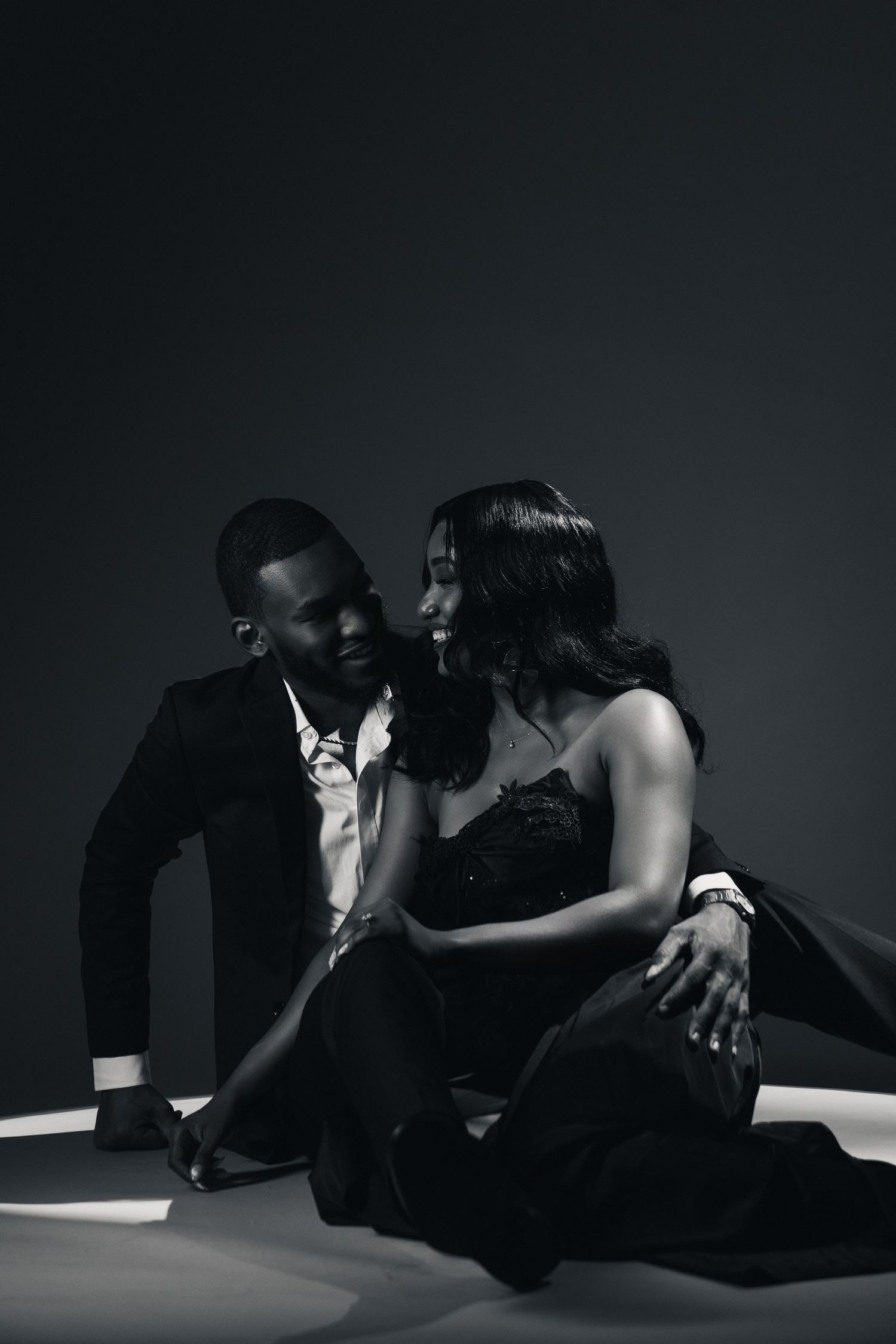 Black and white photo of an elegant couple in formal wear sitting together in a dramatic studio lighting setting.