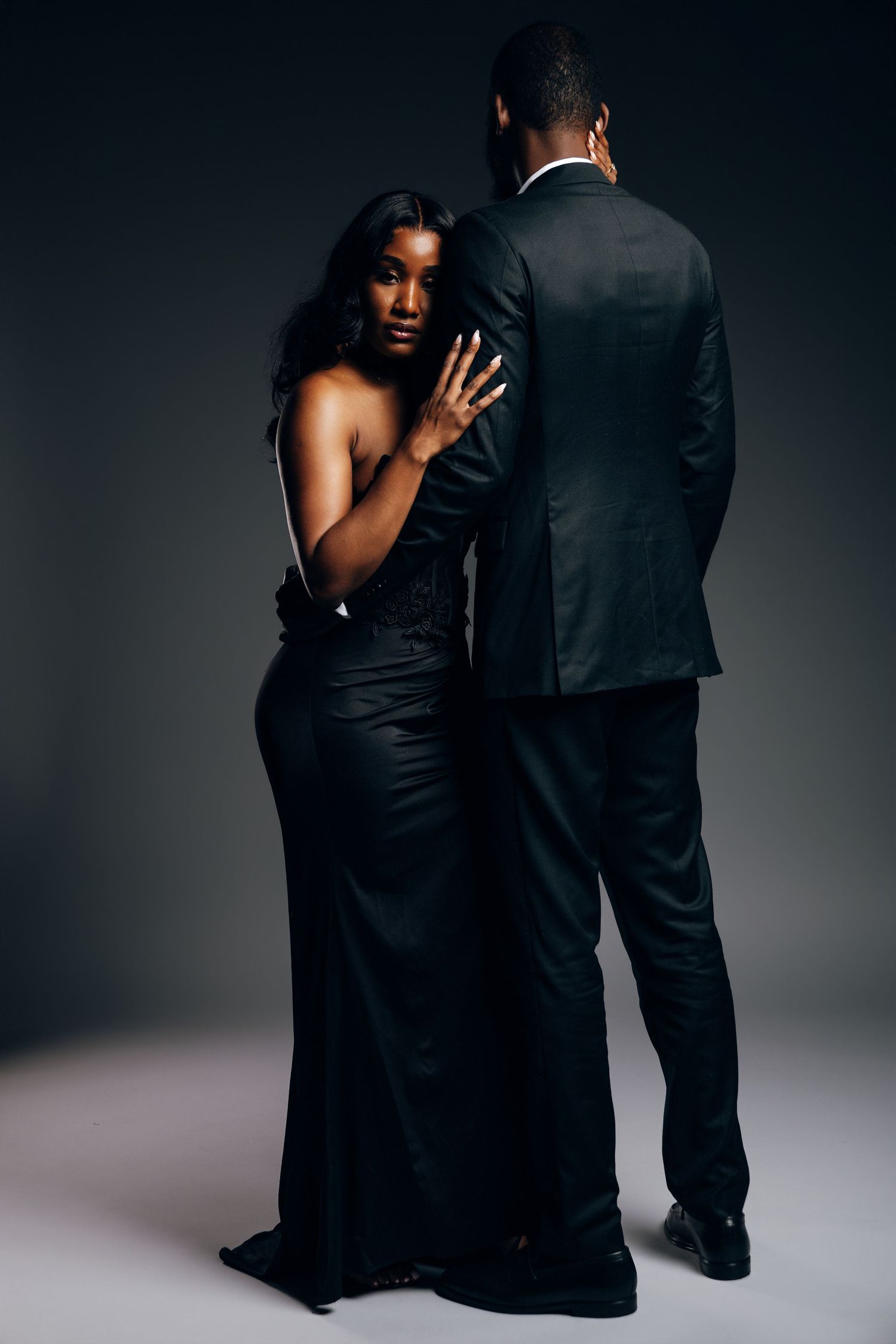 Elegant Black couple in formal attire, woman in gown embraces man in suit, posing together in a dark studio setting.