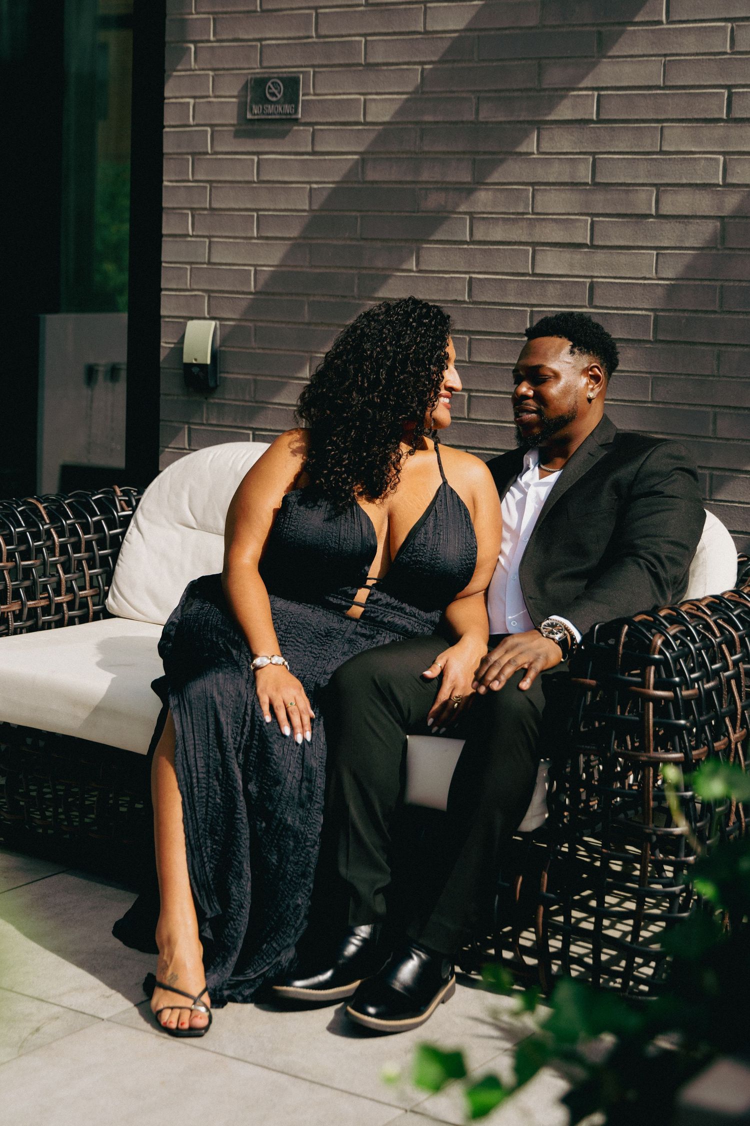 Elegant couple in black formal attire sitting together on a wicker sofa against a gray brick wall.