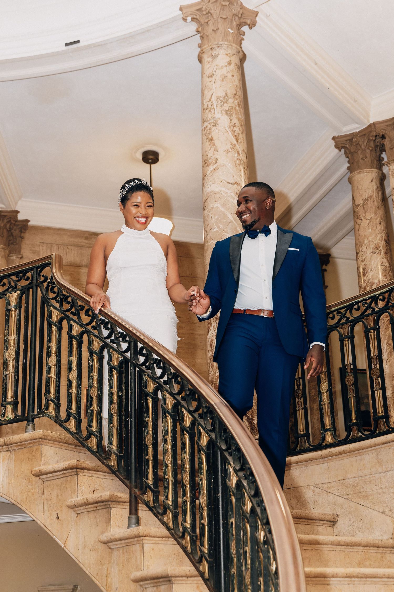 Newlyweds descend elegant curved staircase with ornate railing, groom in blue tuxedo, bride in white gown.
