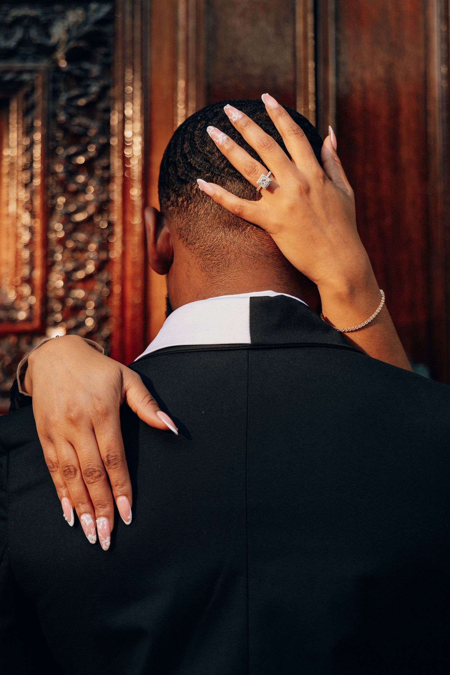 A woman's bejeweled hands embrace a man in a black suit from behind near ornate wooden doors.