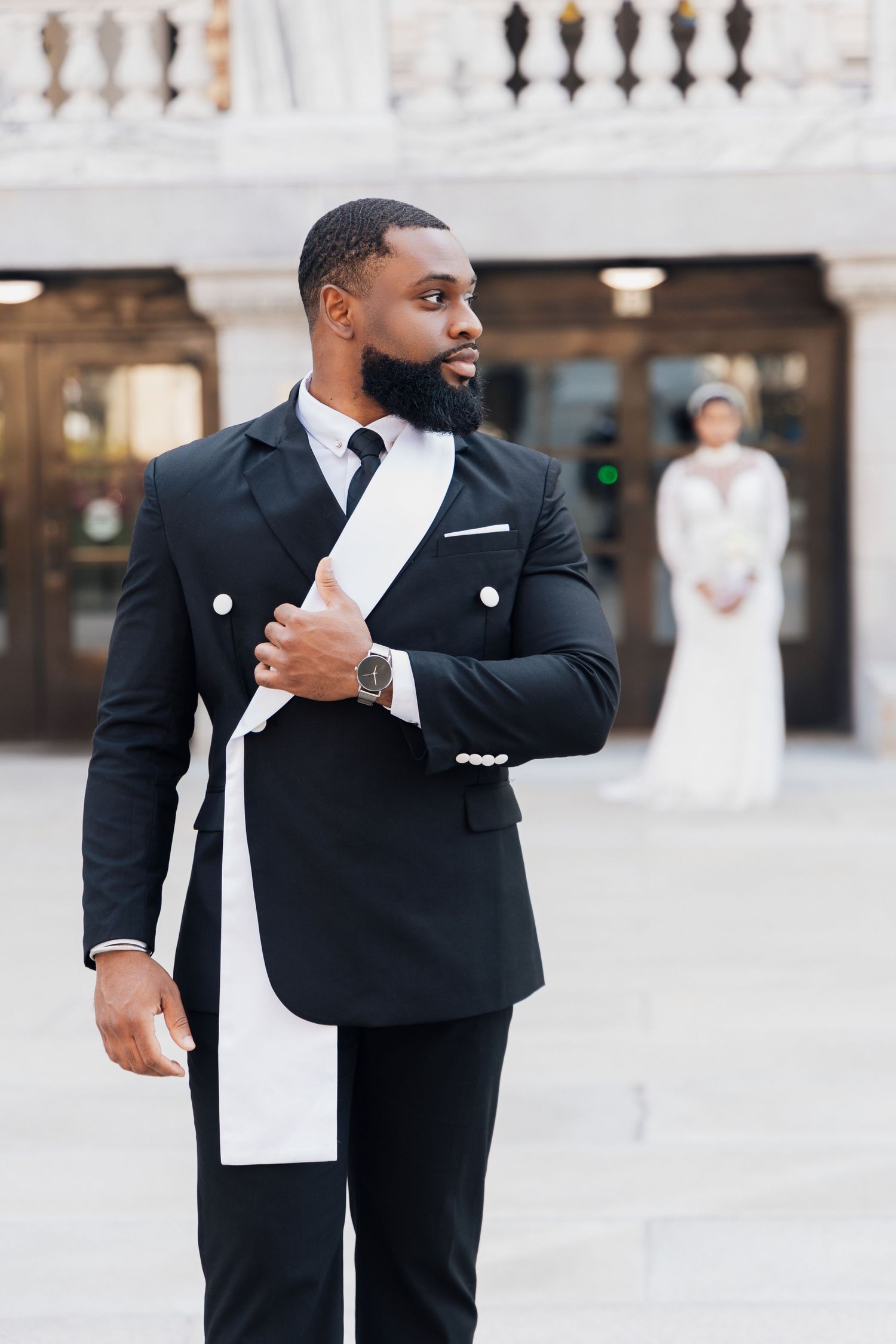 Elegant groom in black double-breasted suit with white tie, standing outdoors with bride in white dress in background.