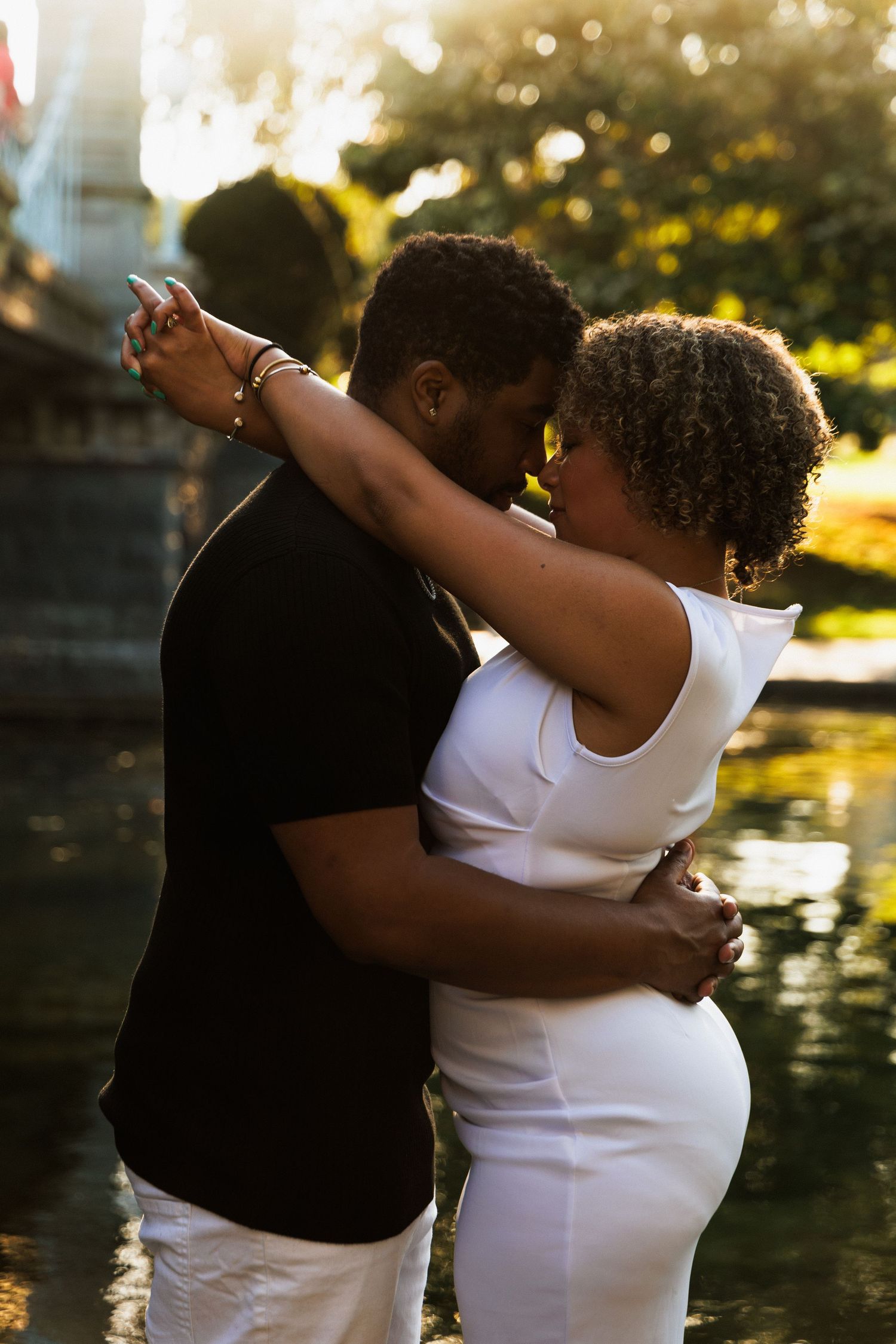 A couple shares a romantic embrace near a sunlit pond in a lush park, she in white and he in black.