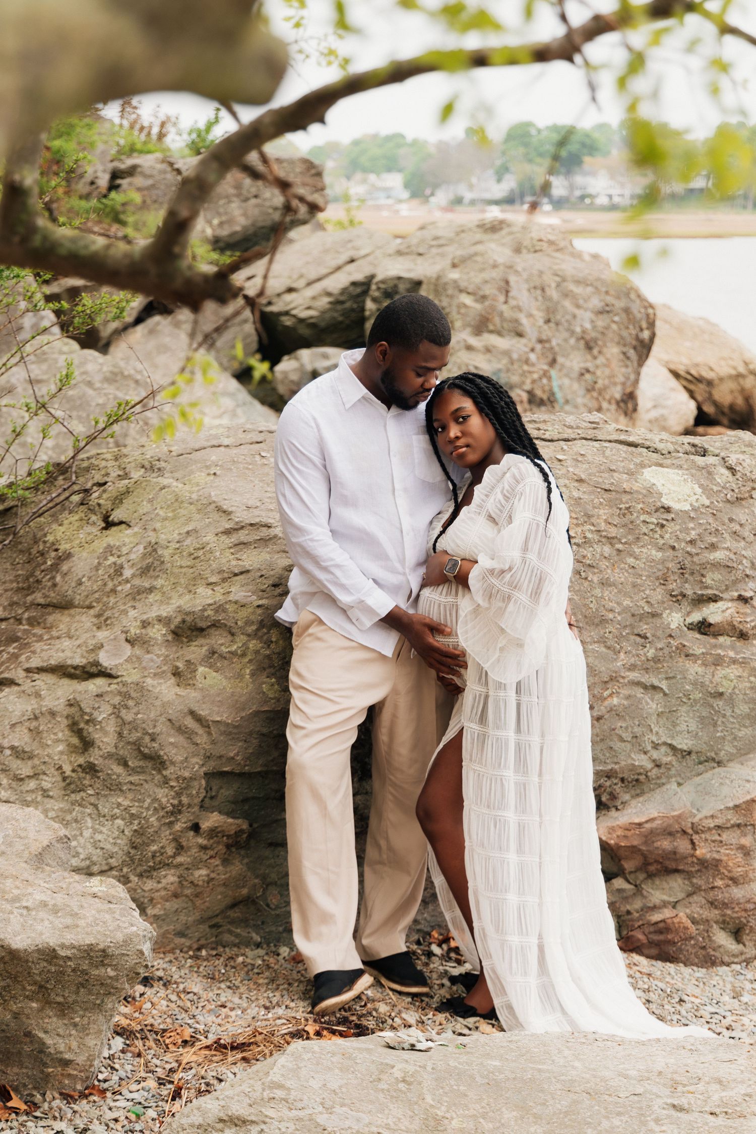 A pregnant couple in white embraces tenderly against large rocks near water.