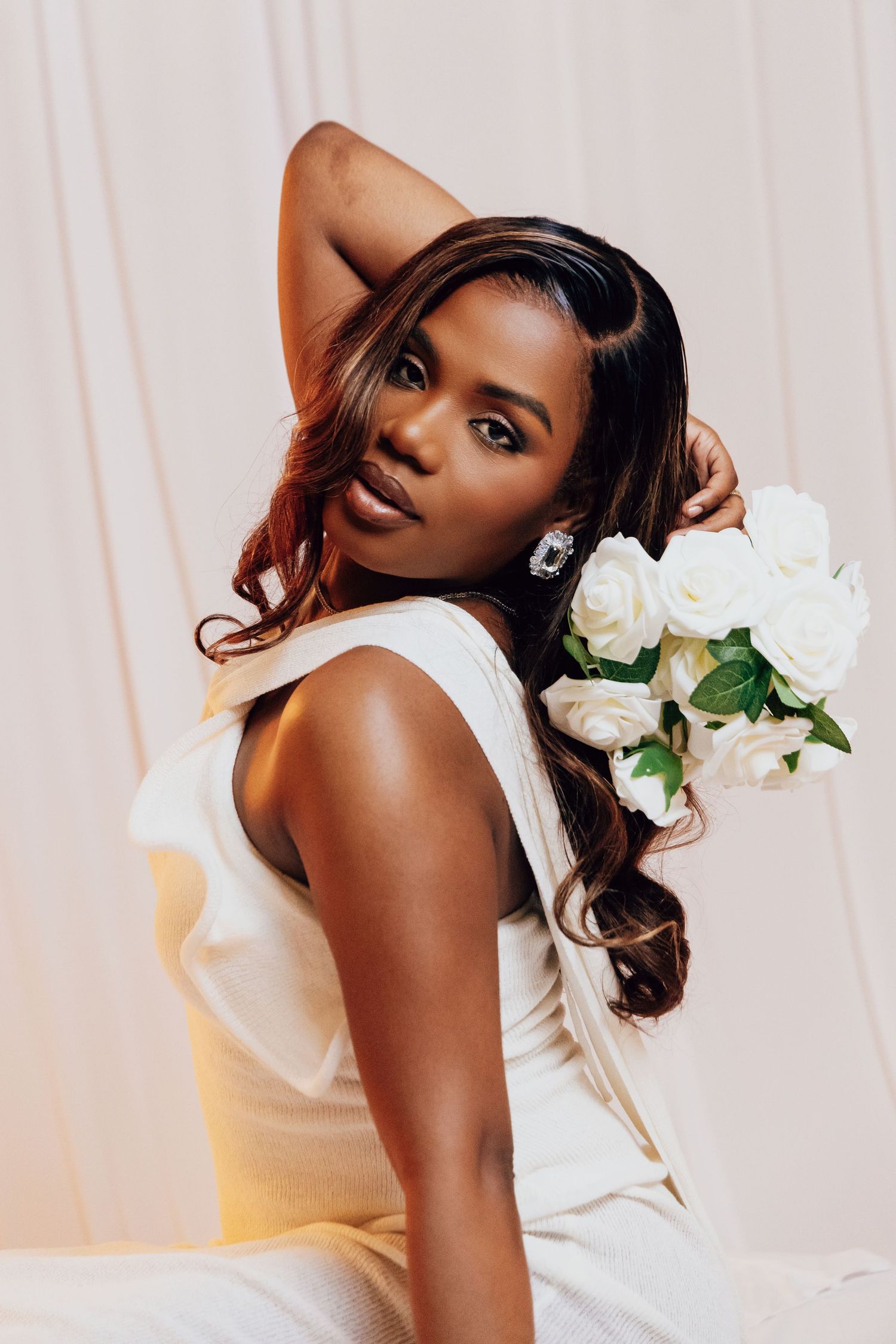 A beautiful Black woman in a white dress holds white roses, posing elegantly with her hand behind her head.