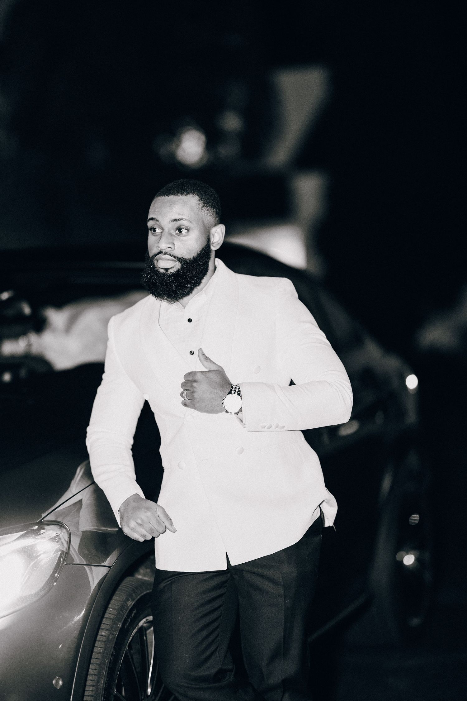 Black and white portrait of bearded groom in white suit jacket posing confidently beside a luxury car.