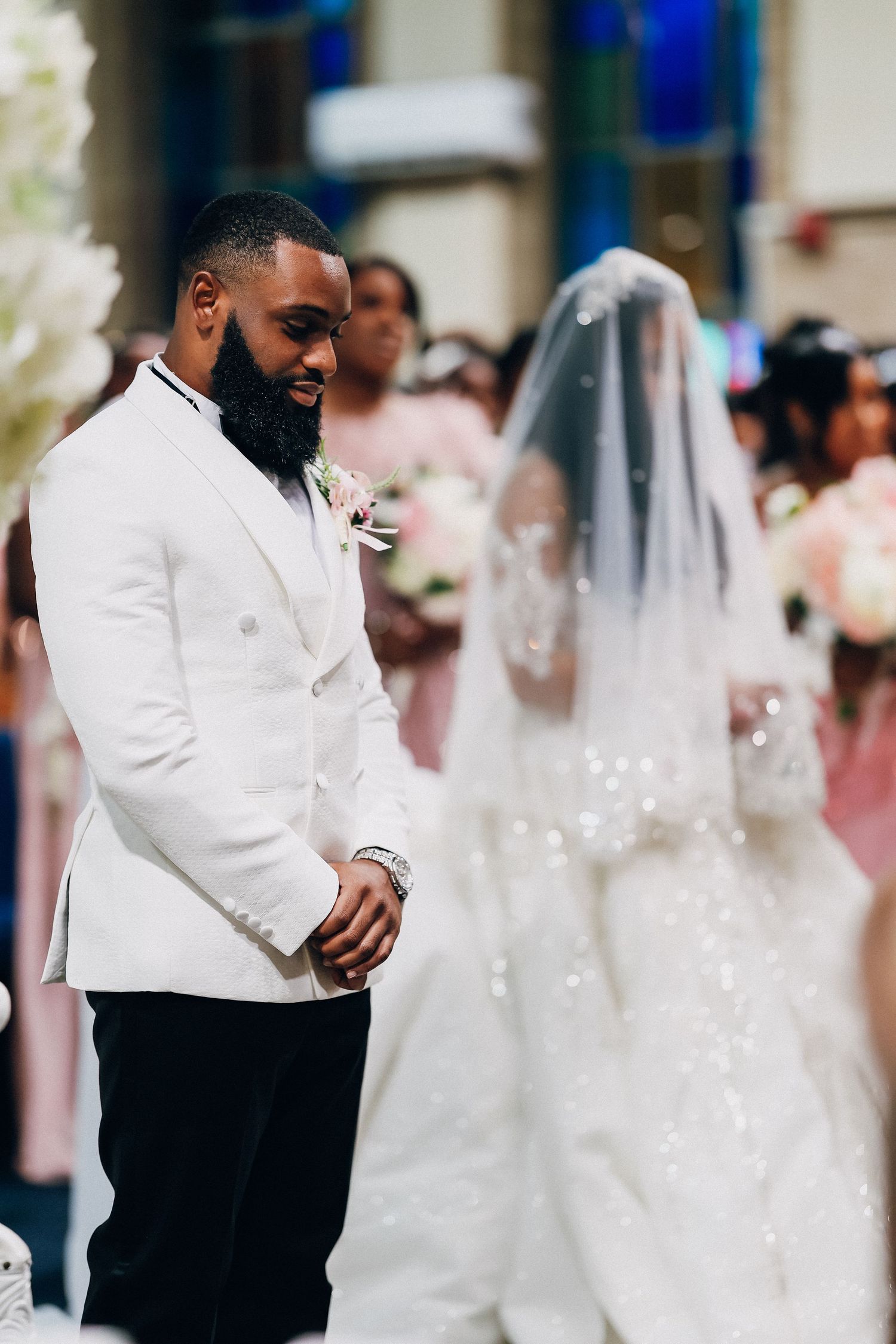 A groom in a white tuxedo smiles at his bride in a beaded wedding gown and veil during their church ceremony.