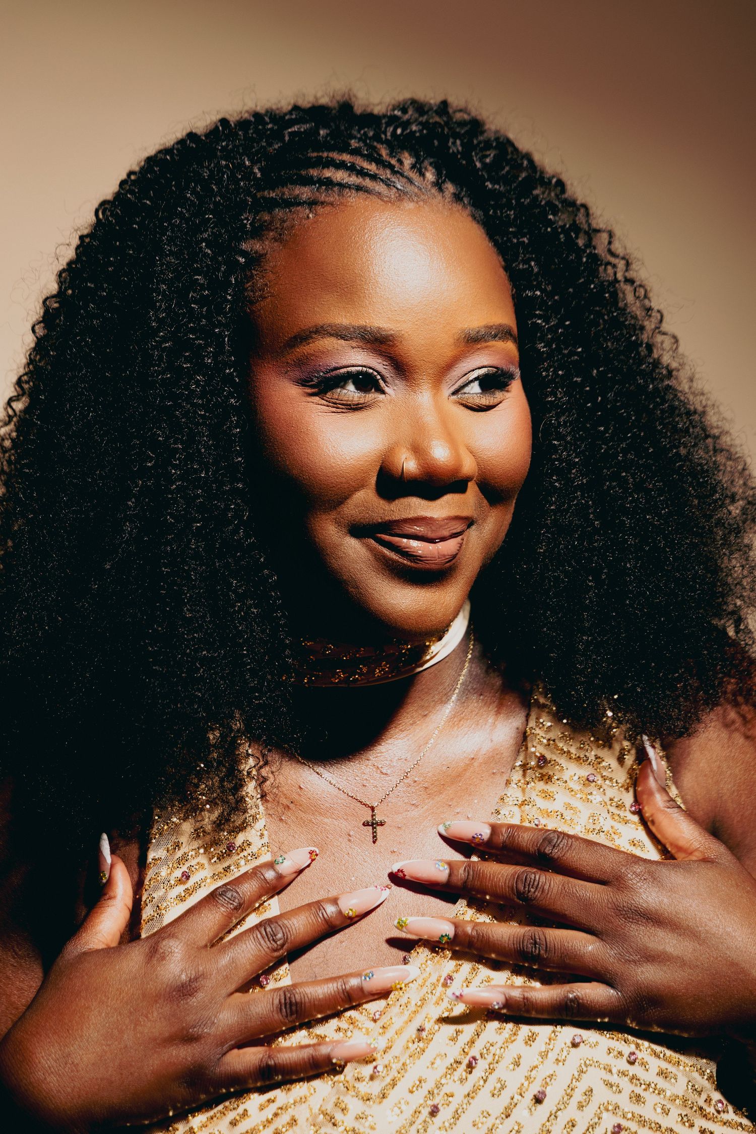 A young Black woman with voluminous curly hair smiles warmly, wearing a gold outfit and cross necklace, hands on chest.