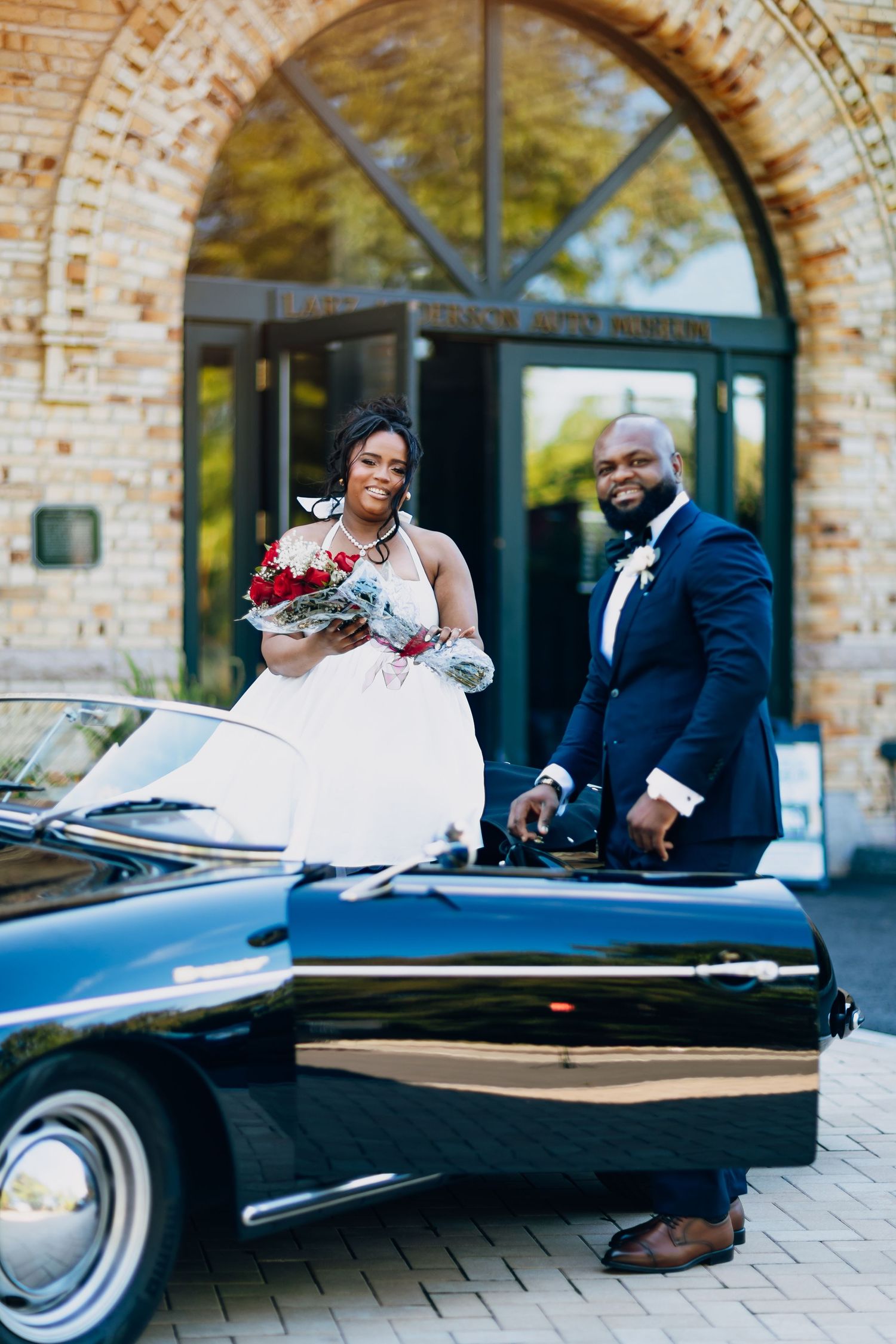 A bride holding a red bouquet and groom pose beside a classic black car at a stone venue.