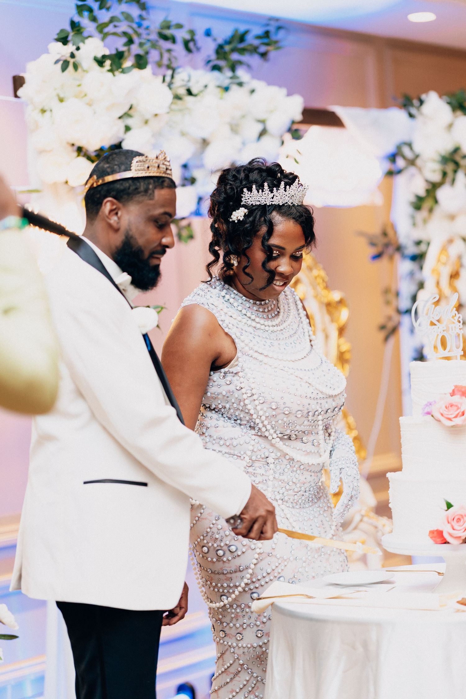 Bride and groom cutting wedding cake, bride wearing crystal gown and tiara, groom in white tuxedo.