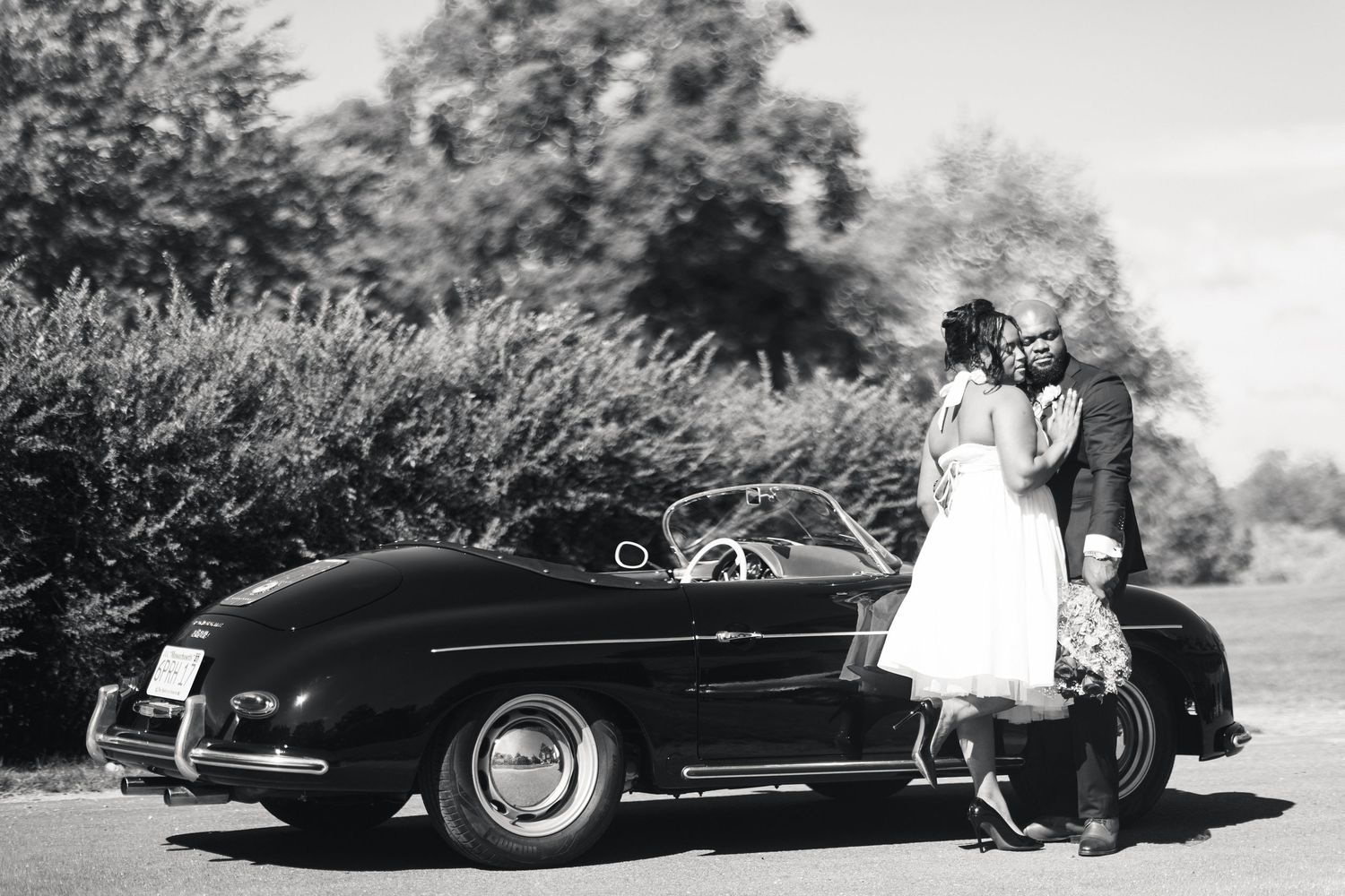 Black and white photo of newlyweds sharing a kiss beside a classic vintage convertible surrounded by lush greenery.