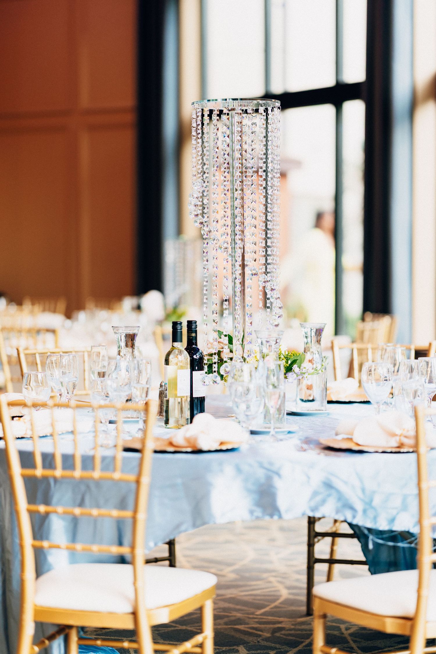 Formal wedding reception tables with blue linens, gold chiavari chairs, crystal chandelier centerpiece, and wine bottles.