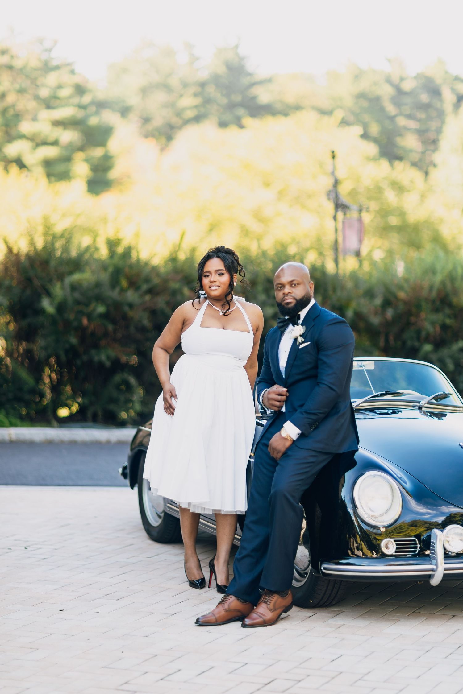 Bride in white halter dress and groom in navy suit pose beside a vintage black Porsche outdoors.