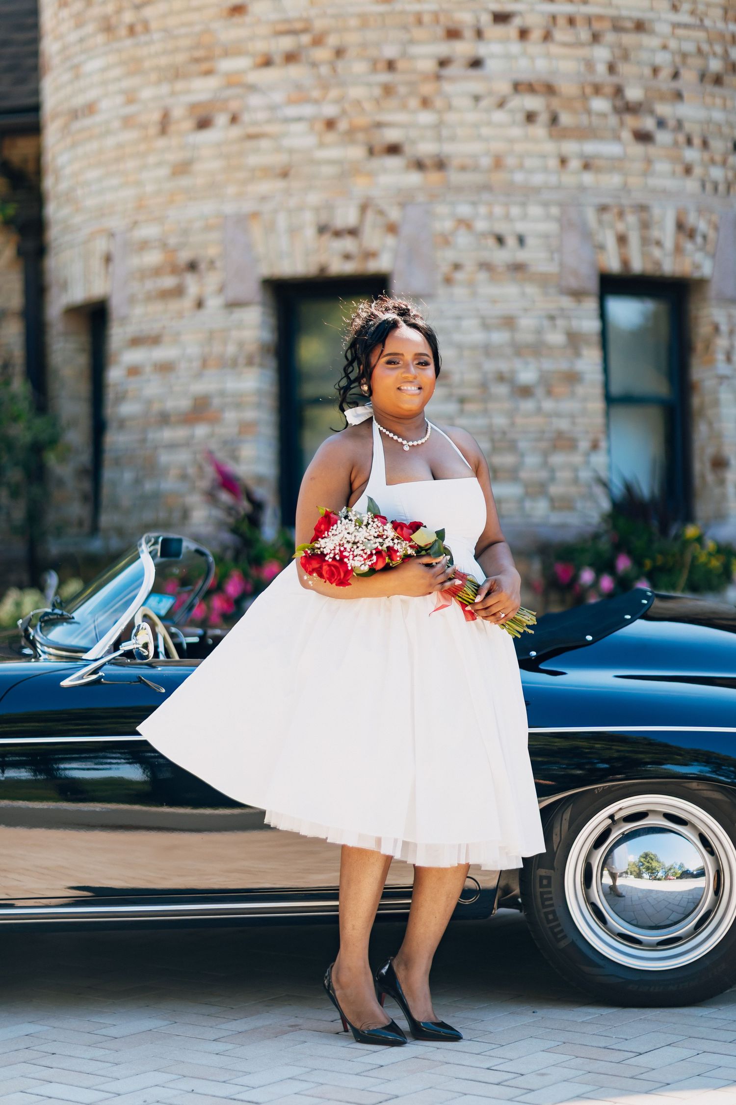 Black bride in white tea-length dress holding floral bouquet poses beside vintage classic car outside brick building.