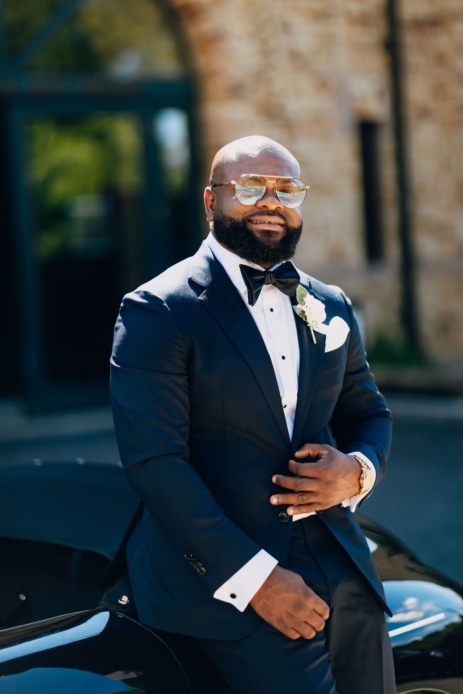 Handsome groom in navy tuxedo with boutonniere and gold aviator sunglasses poses by vintage car.