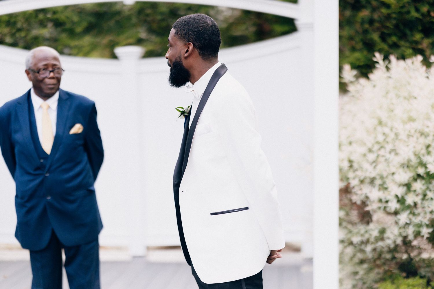 Groom in white tuxedo jacket with black lapels stands outdoors at wedding ceremony with elder man in navy suit.