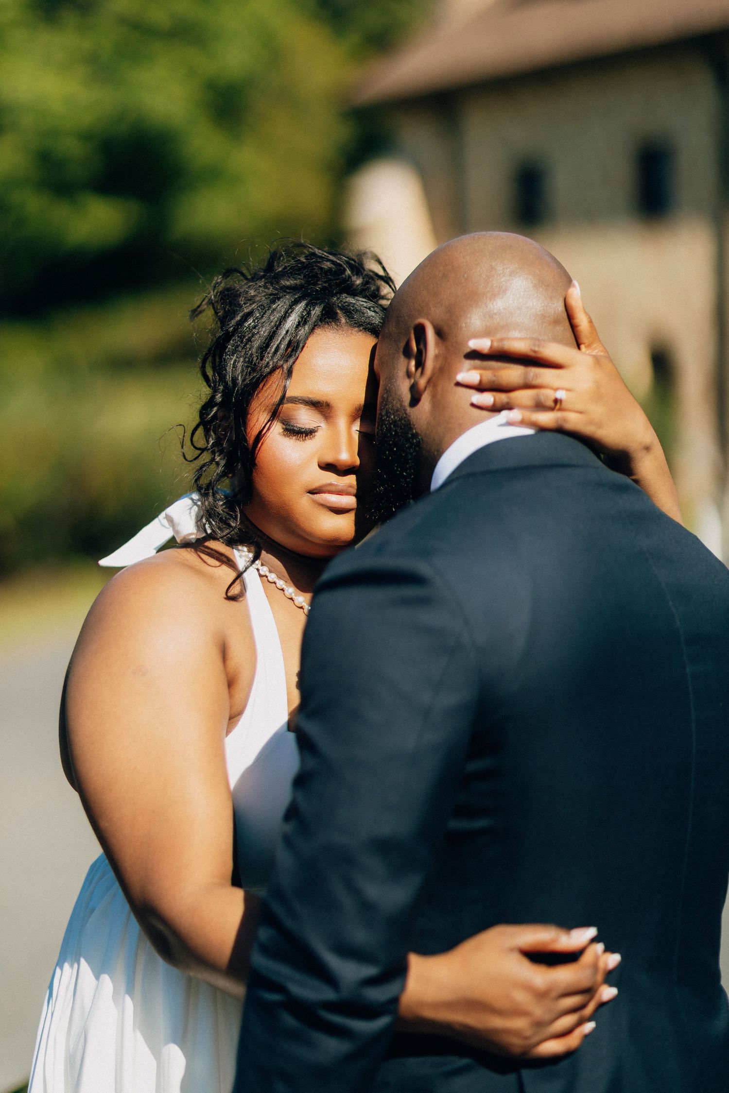 Bride with closed eyes embraces groom tenderly near a stone building on their wedding day.