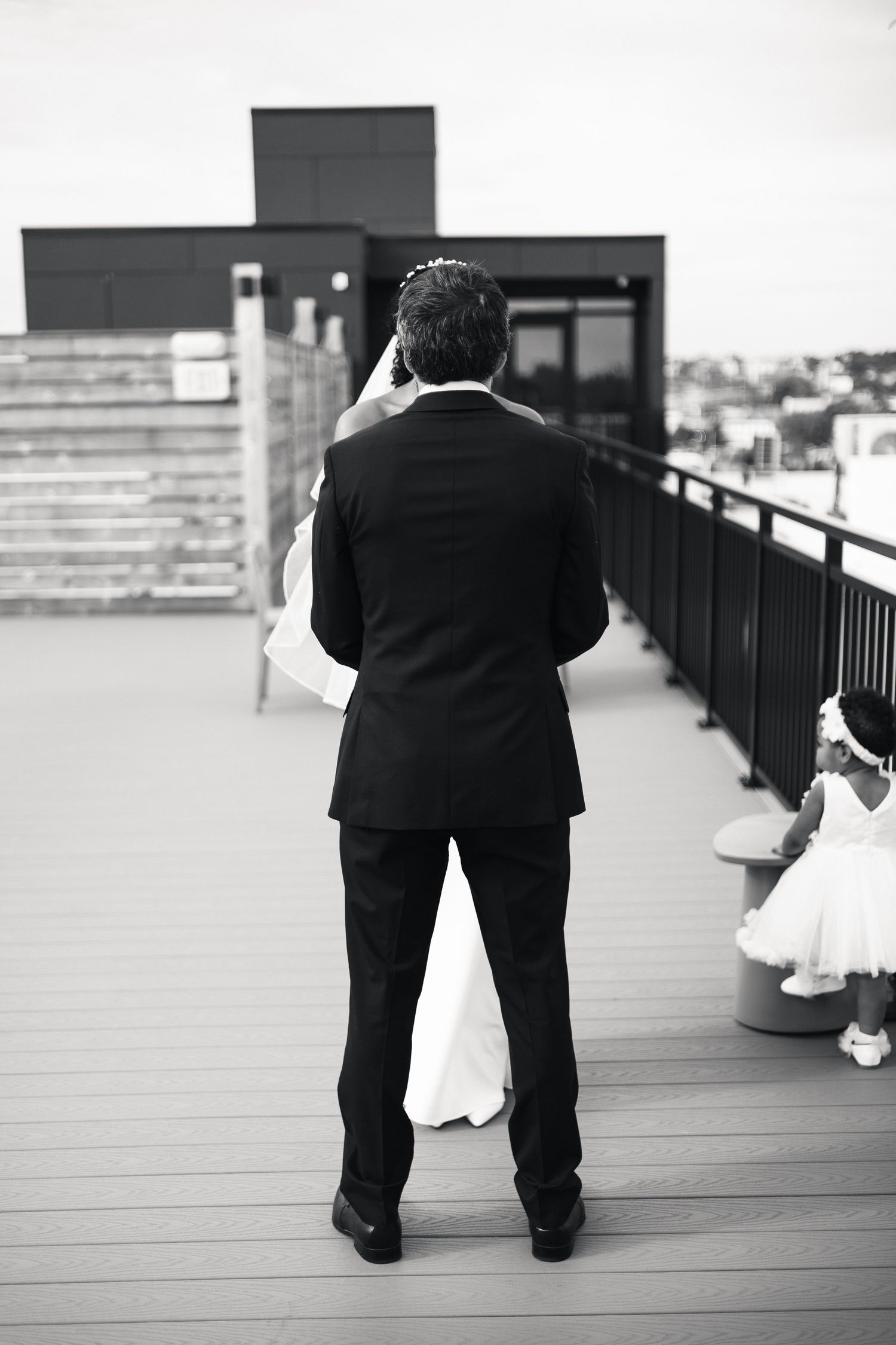 Groom in black suit stands on rooftop deck facing bride in white wedding dress during first look moment.