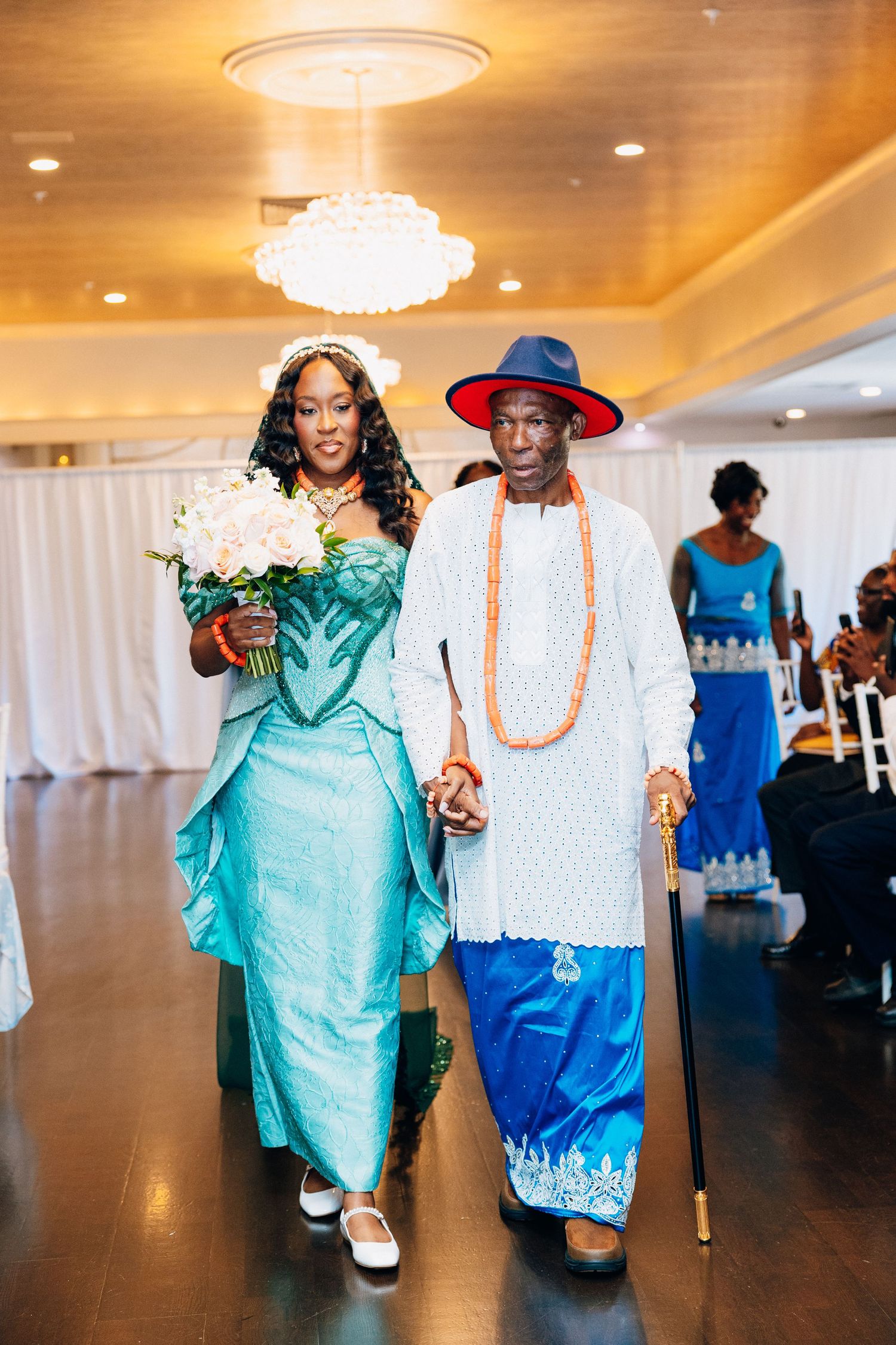 Bride in teal traditional African gown walks down aisle with elder man in white embroidered attire and red hat.