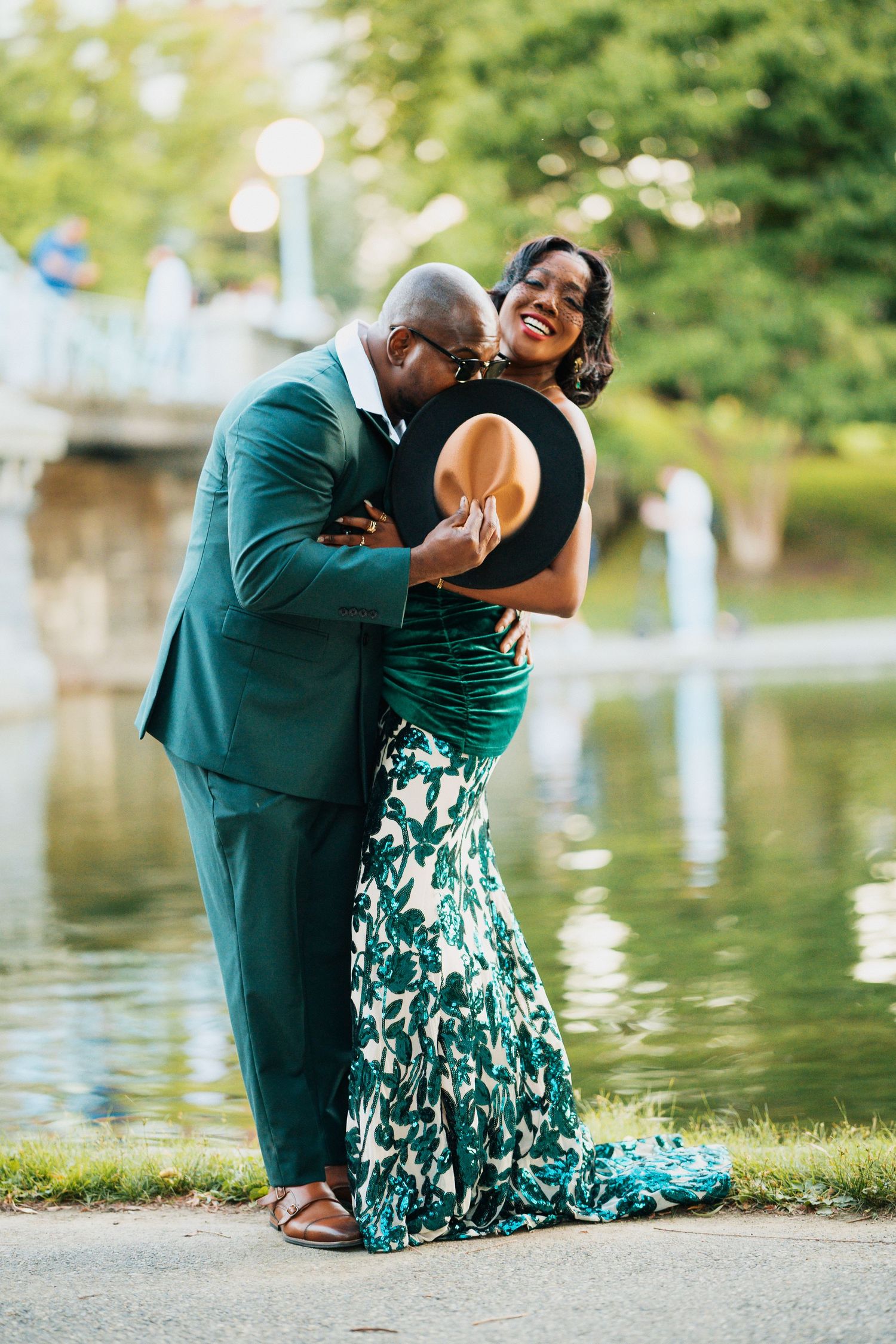 Joyful Black couple embracing by a pond, both wearing coordinated green outfits, woman laughing in floral gown.
