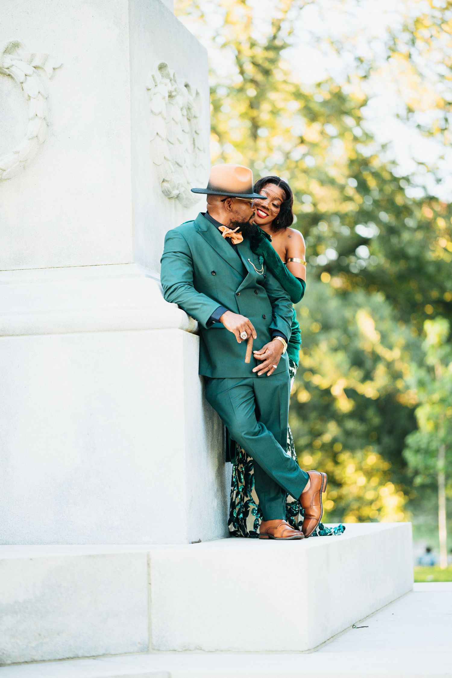 Couple in matching green outfits posing romantically against a white stone column in a sunlit park.