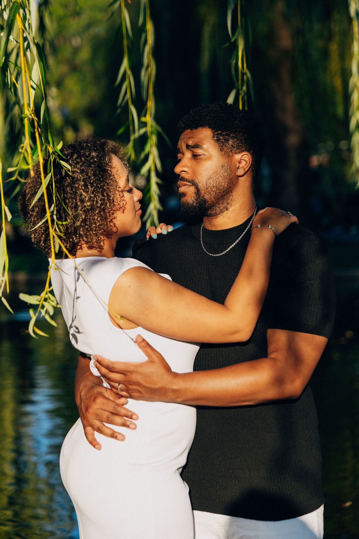 A loving Black couple embrace near a serene pond under weeping willow branches in warm sunlight.