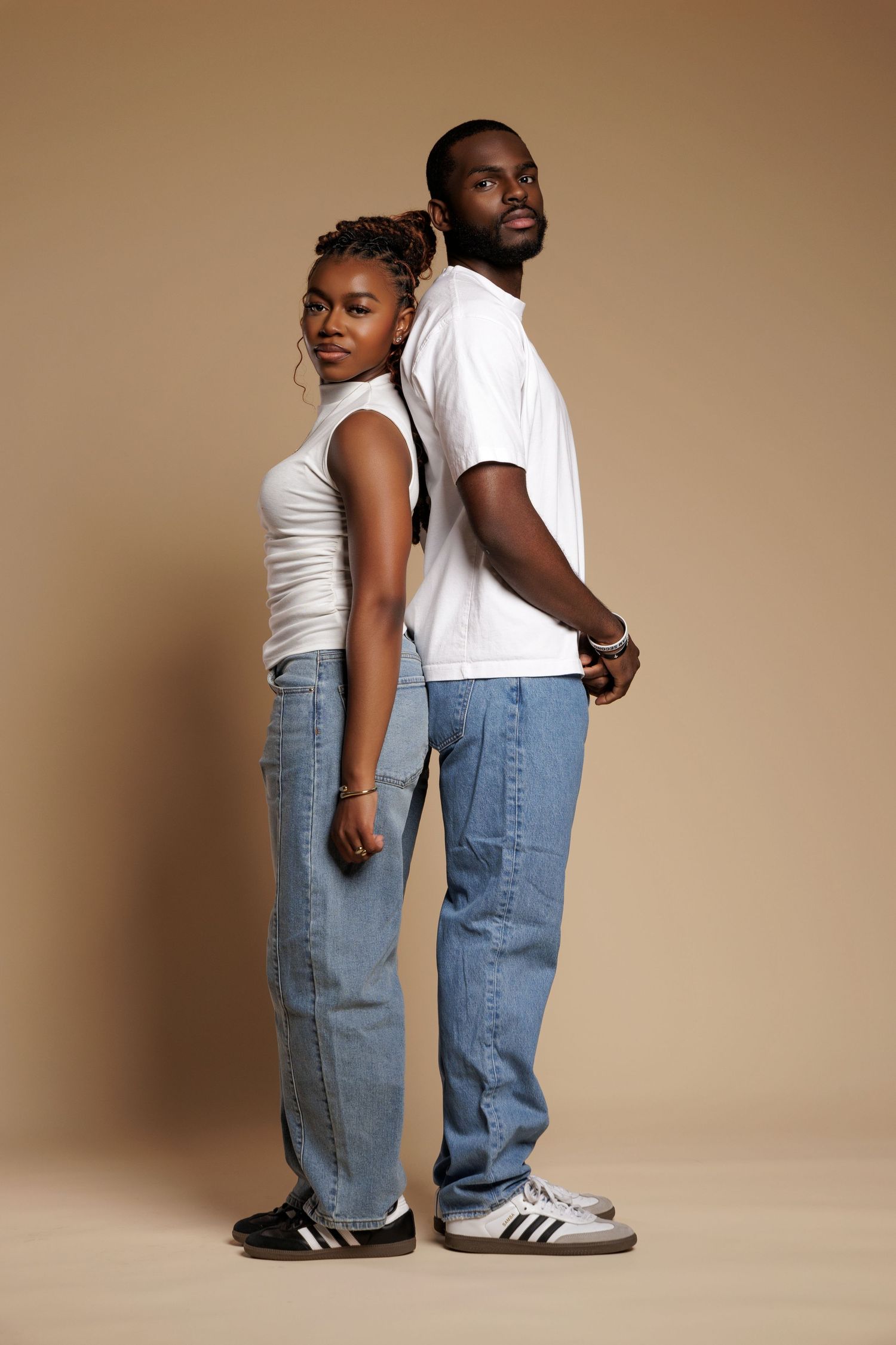 A Black couple in matching white tops and blue jeans pose back-to-back against a beige studio background.