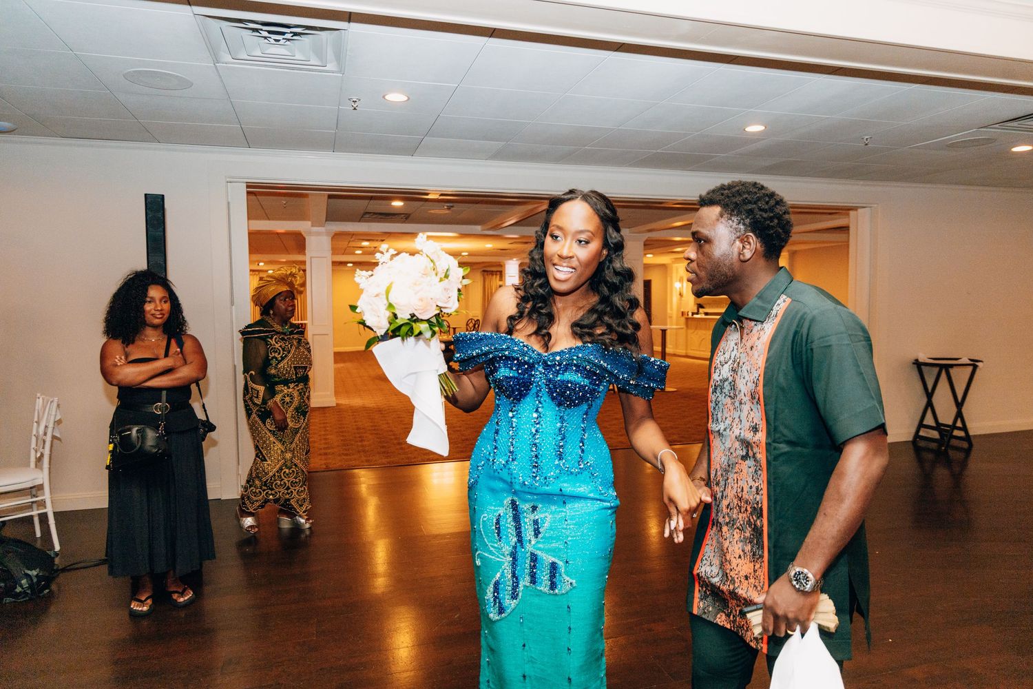 Bride in stunning blue sequin gown holding white bouquet walks with groom in green at wedding reception.