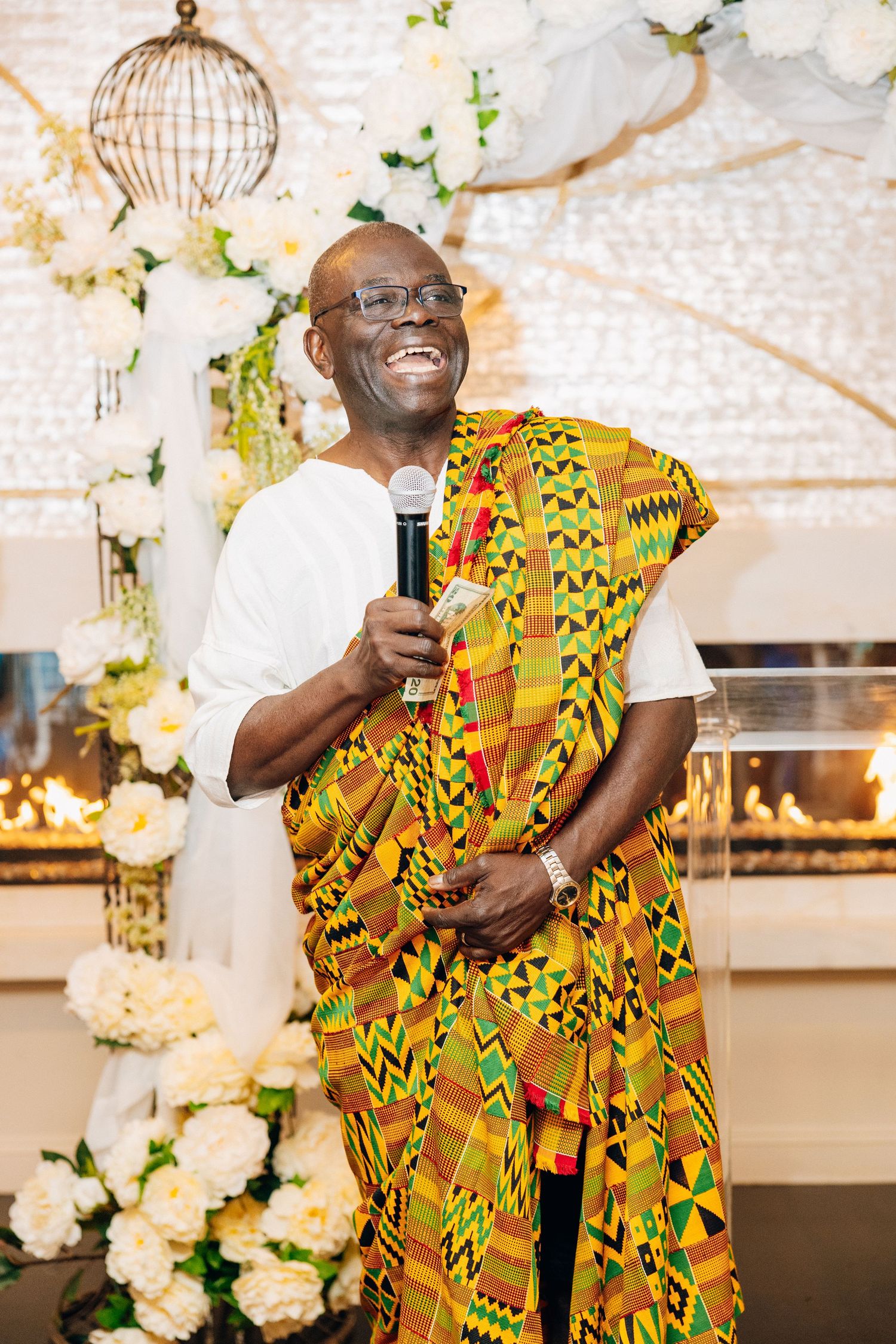 Joyful man in colorful kente cloth laughing while holding microphone at wedding event.