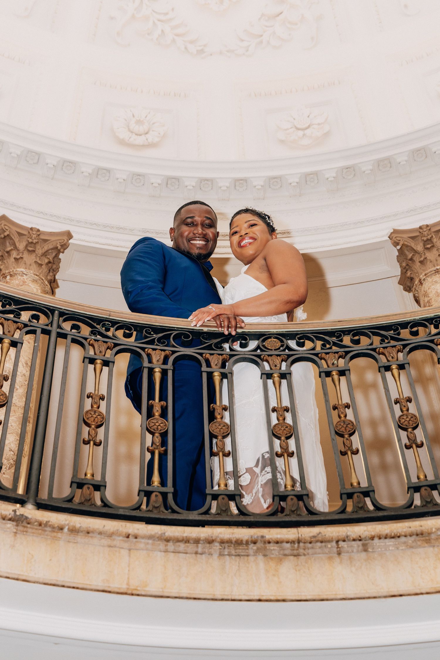 Smiling bride and groom in blue suit lean over ornate iron balcony railing in elegant white domed venue.