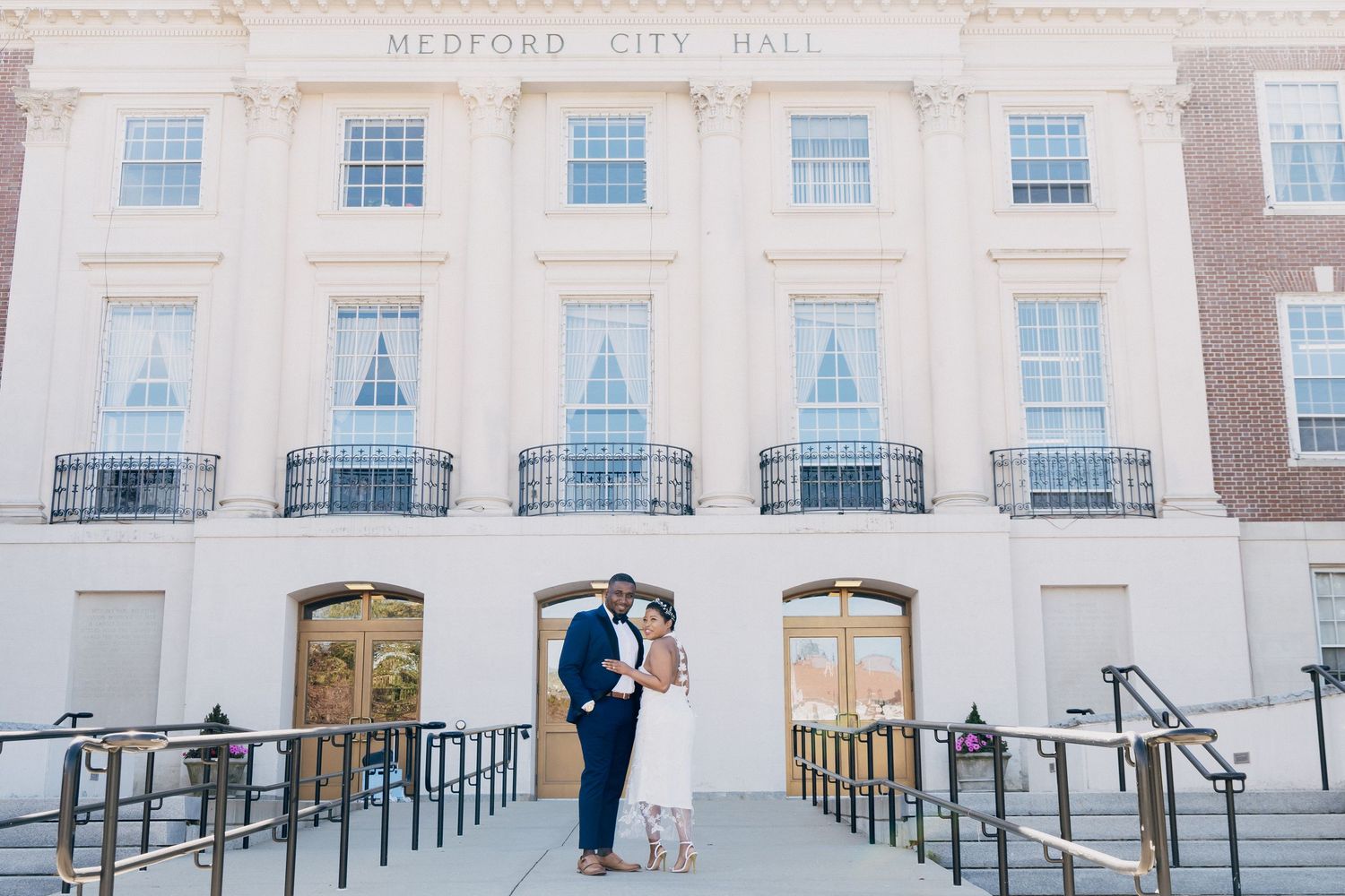 Couple embracing in front of Medford City Hall, bride in floral dress and groom in navy suit.