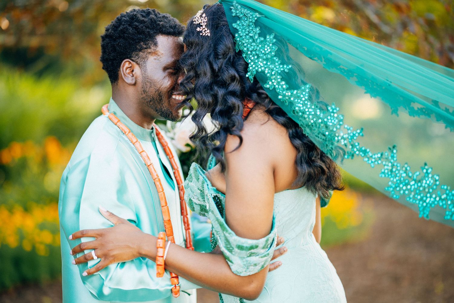 Nigerian bride and groom in coordinating teal traditional outfits embrace among yellow flowers outdoors.