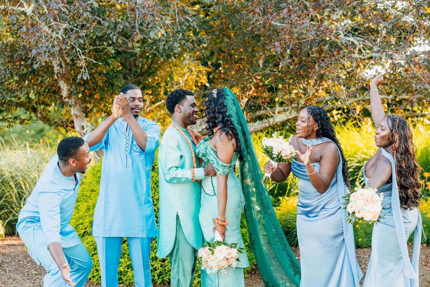 Wedding party celebrates in teal attire outdoors among trees.
