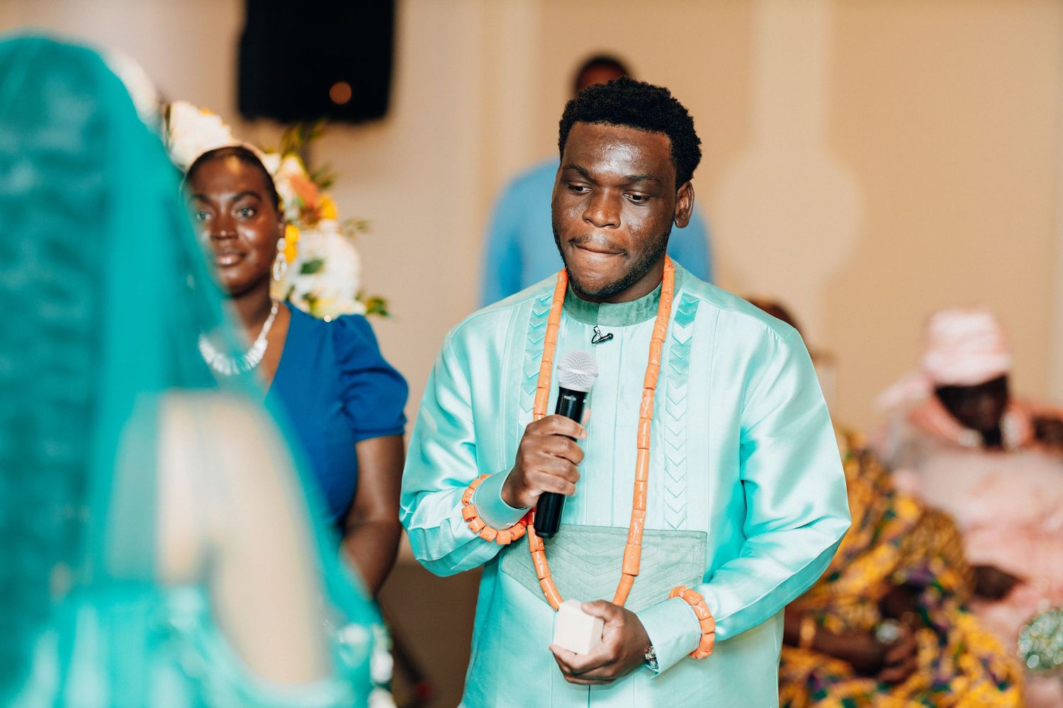 Man in teal African traditional attire holding microphone at a cultural celebration event.