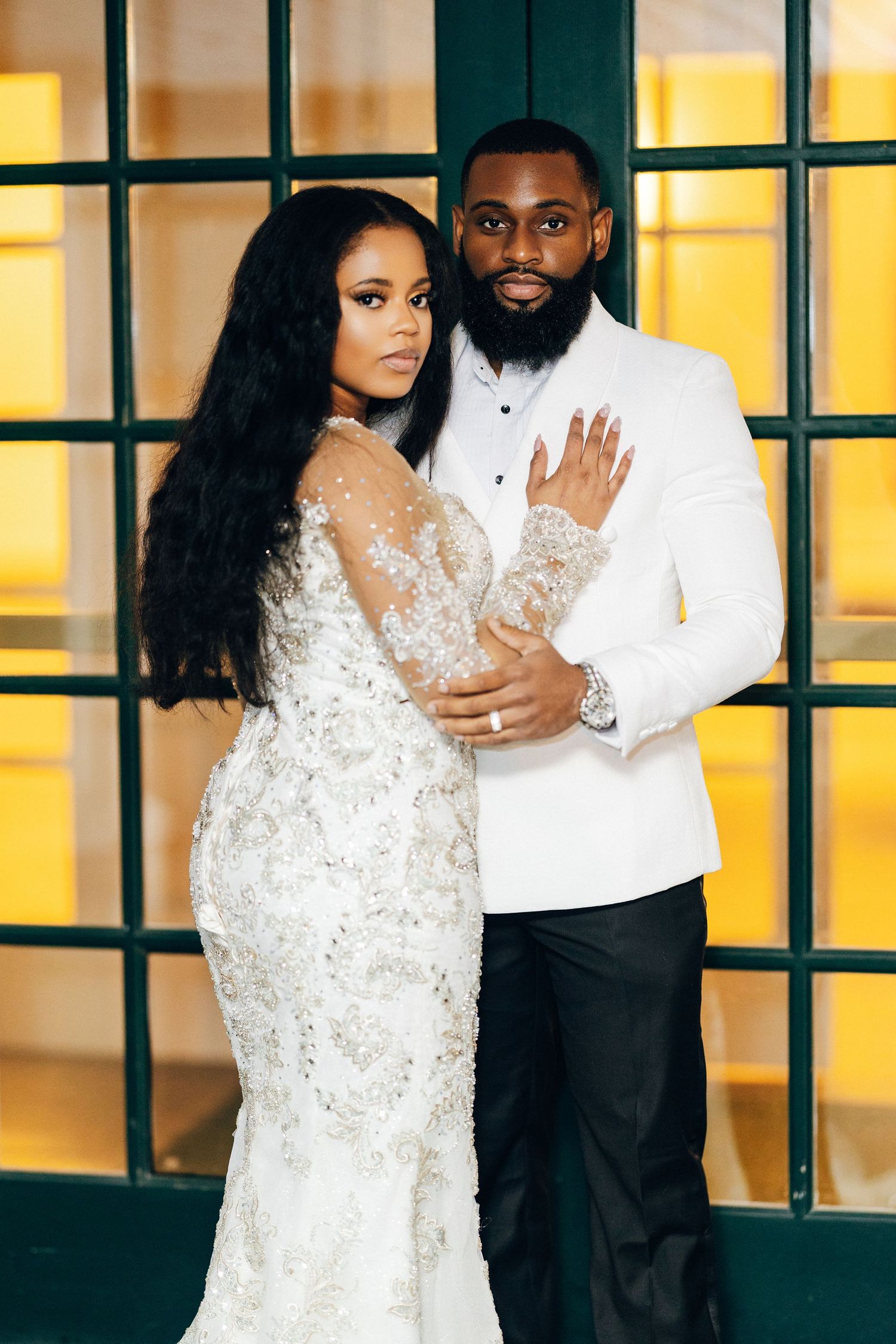 Elegant Black couple posing in wedding attire, bride in beaded gown, groom in white tuxedo jacket.