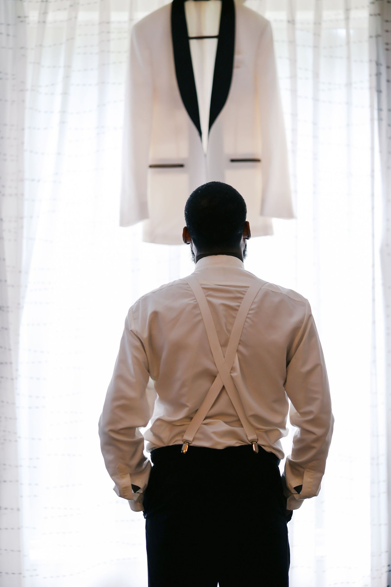 A groom in suspenders gazes at his white tuxedo jacket with black lapels hanging by a bright window.