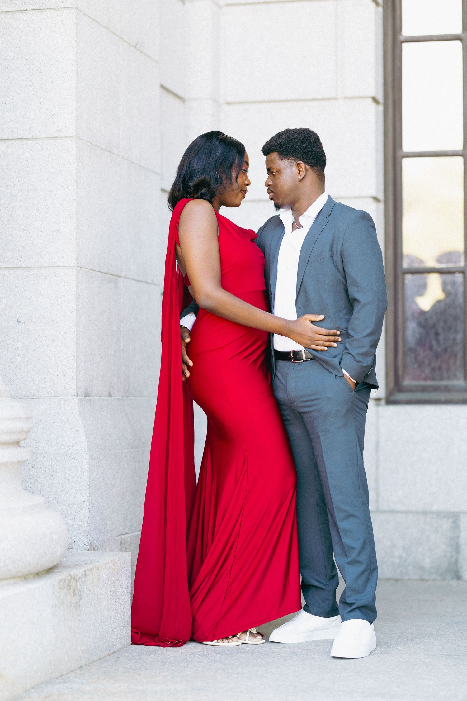 Elegant couple posing outdoors, woman in red cape gown and man in gray suit against stone columns.