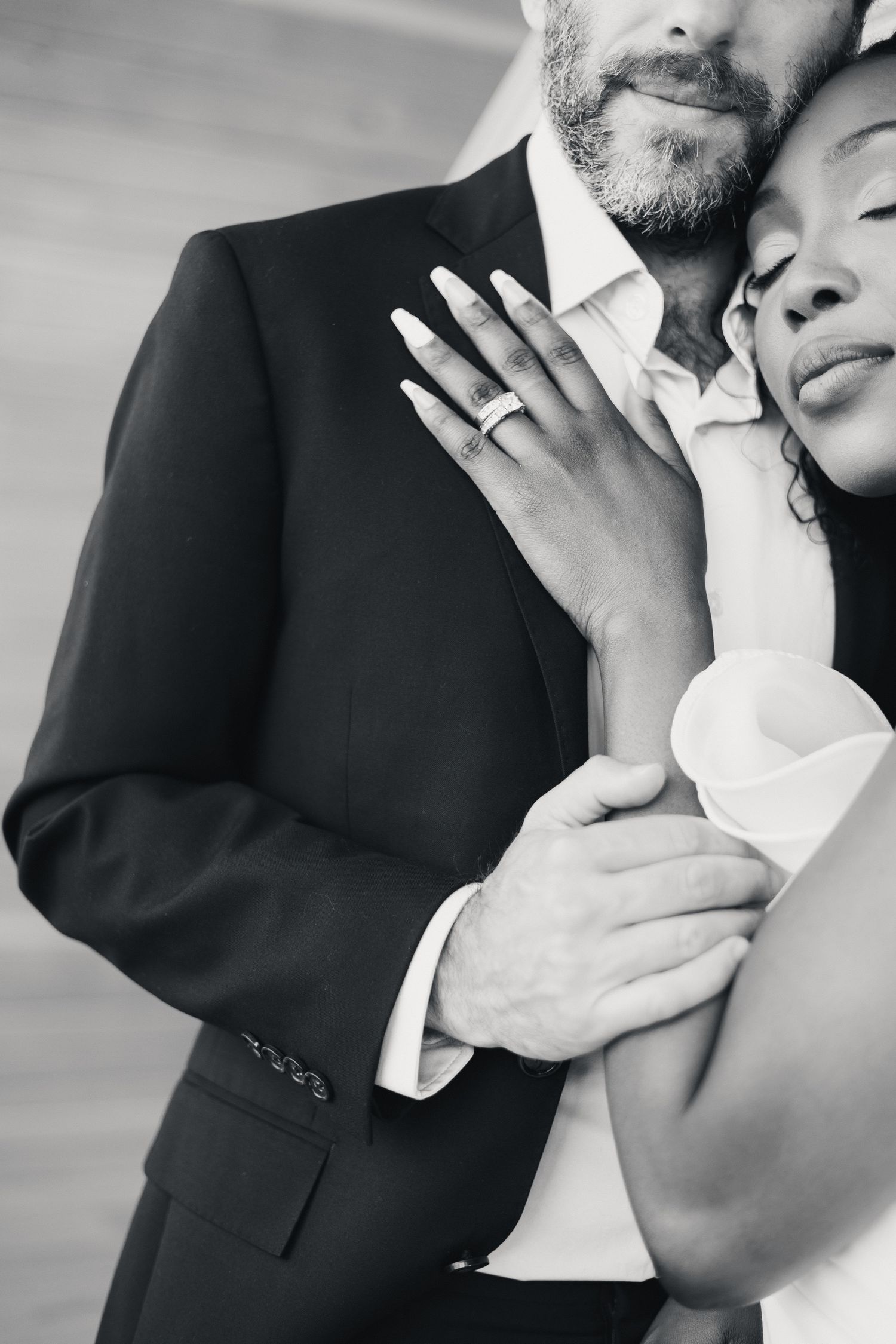 A black and white photo of an interracial couple embracing, showcasing the bride's wedding ring.