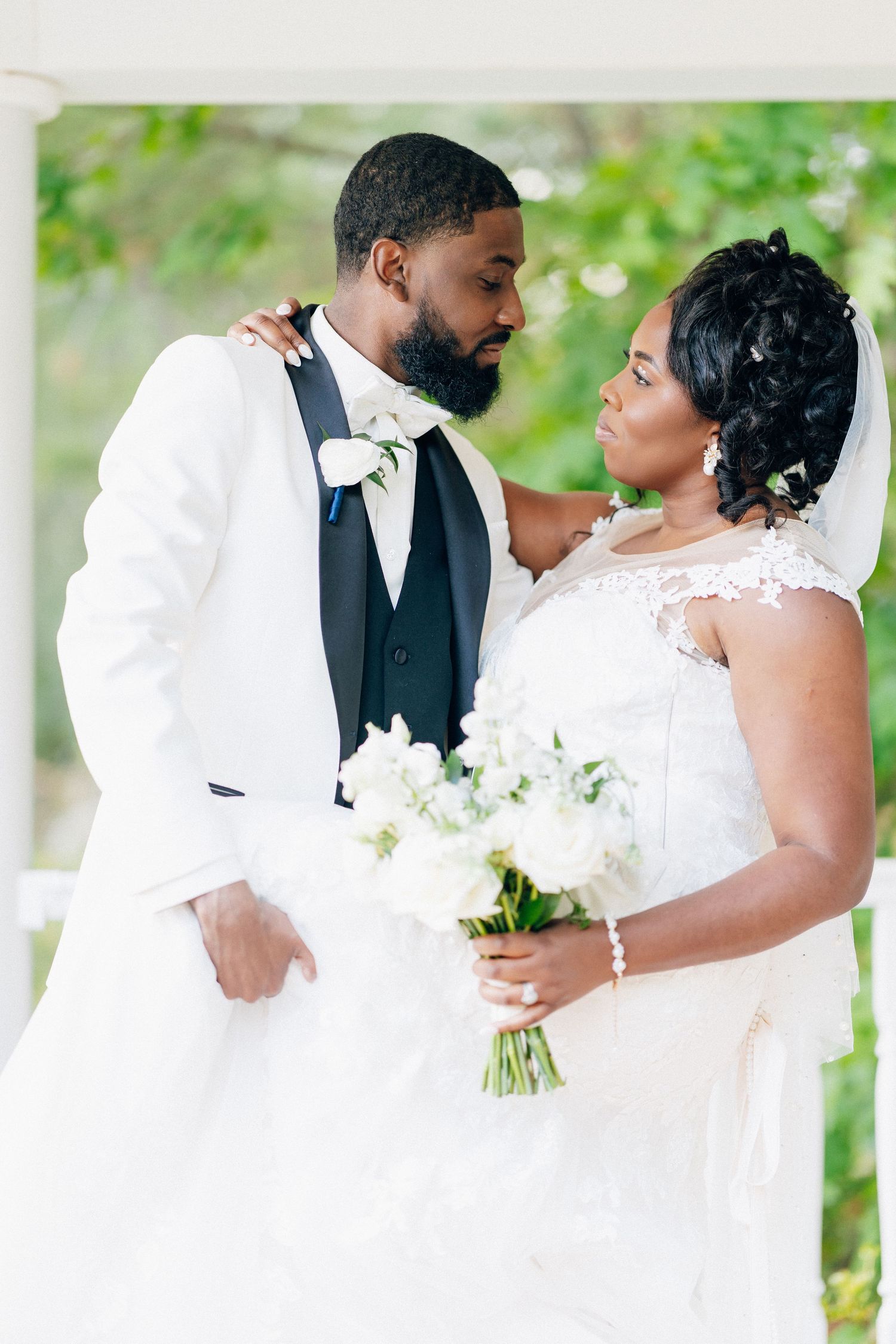A bride and groom in white wedding attire share a tender moment, bride holding white floral bouquet outdoors.