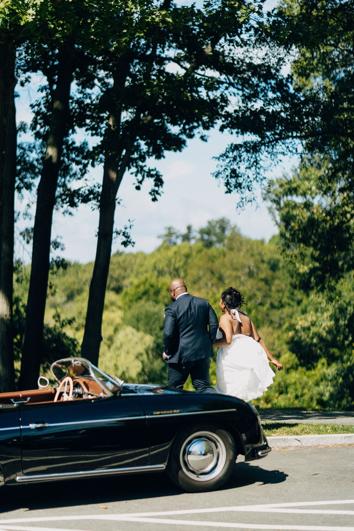 Newlywed couple walking hand in hand past a vintage black convertible toward lush green trees.