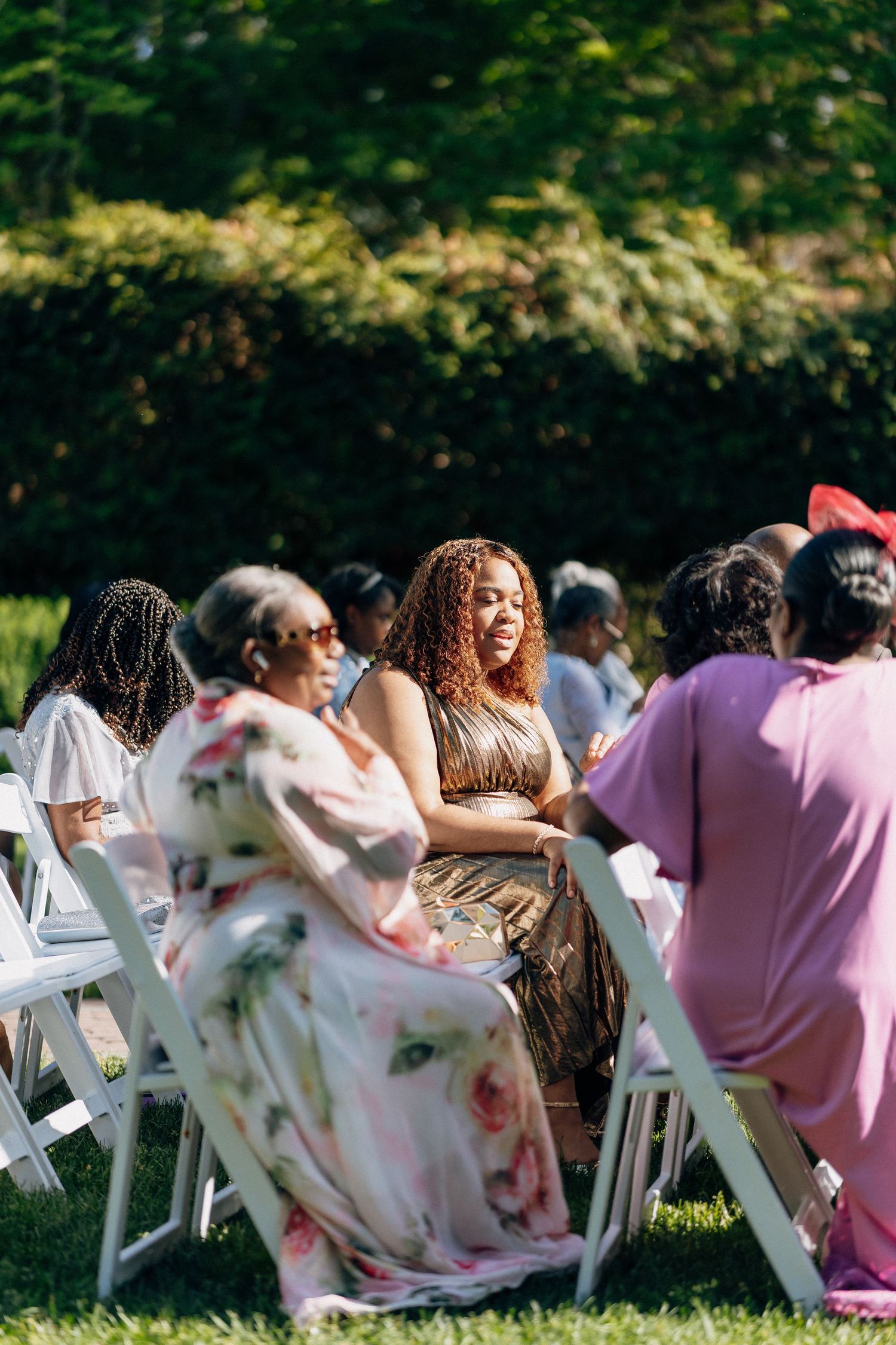 Smiling wedding guests in colorful floral and pink dresses seated on white chairs at an outdoor ceremony.