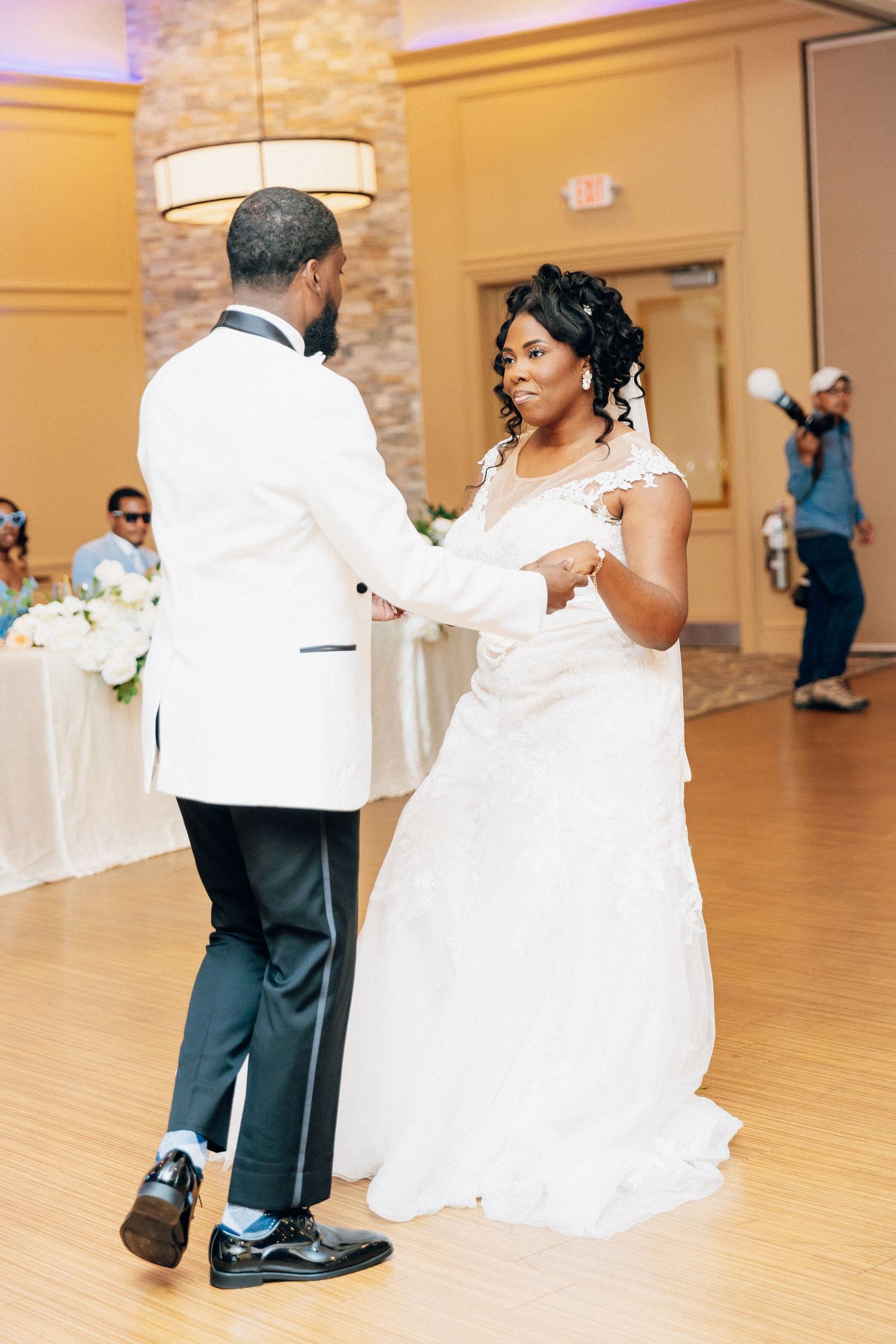 Bride and groom share their first dance at wedding reception, bride wearing lace gown, groom in white tuxedo jacket.