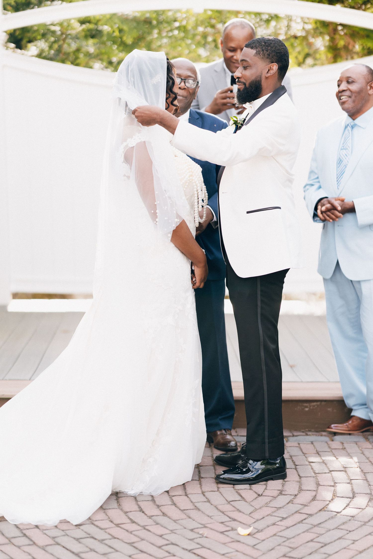 Groom in white tuxedo lifting bride's veil at outdoor wedding ceremony under white pergola.