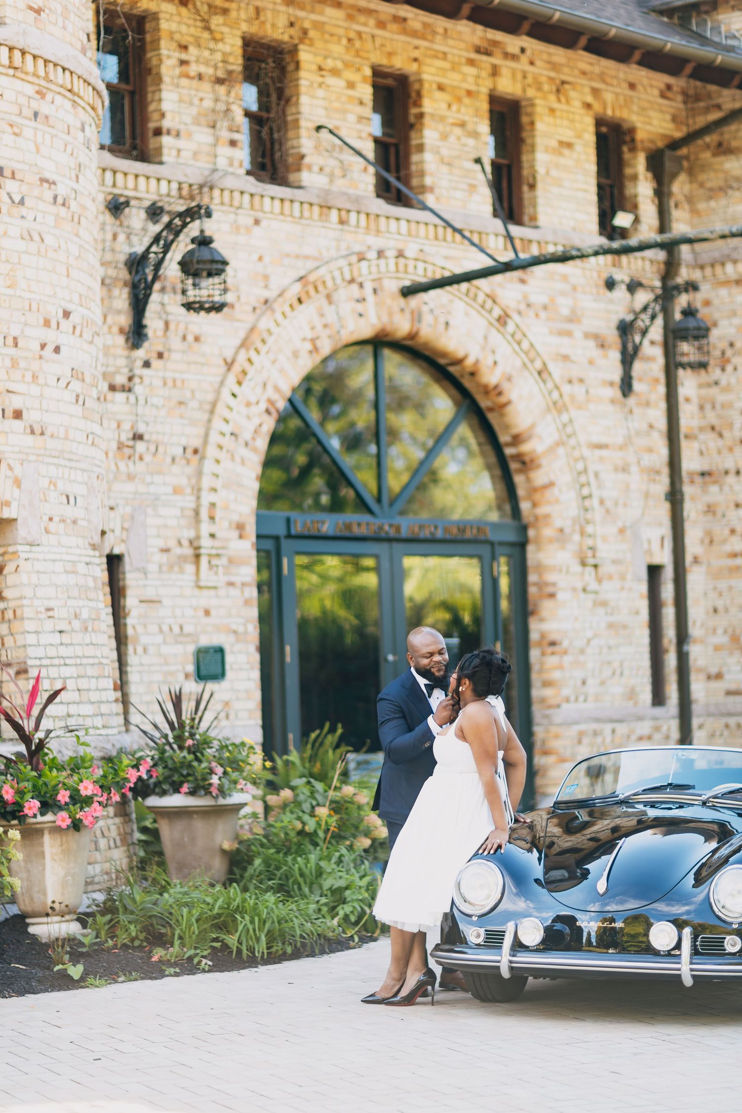 Black couple in wedding attire embracing near a vintage car outside a historic brick building.