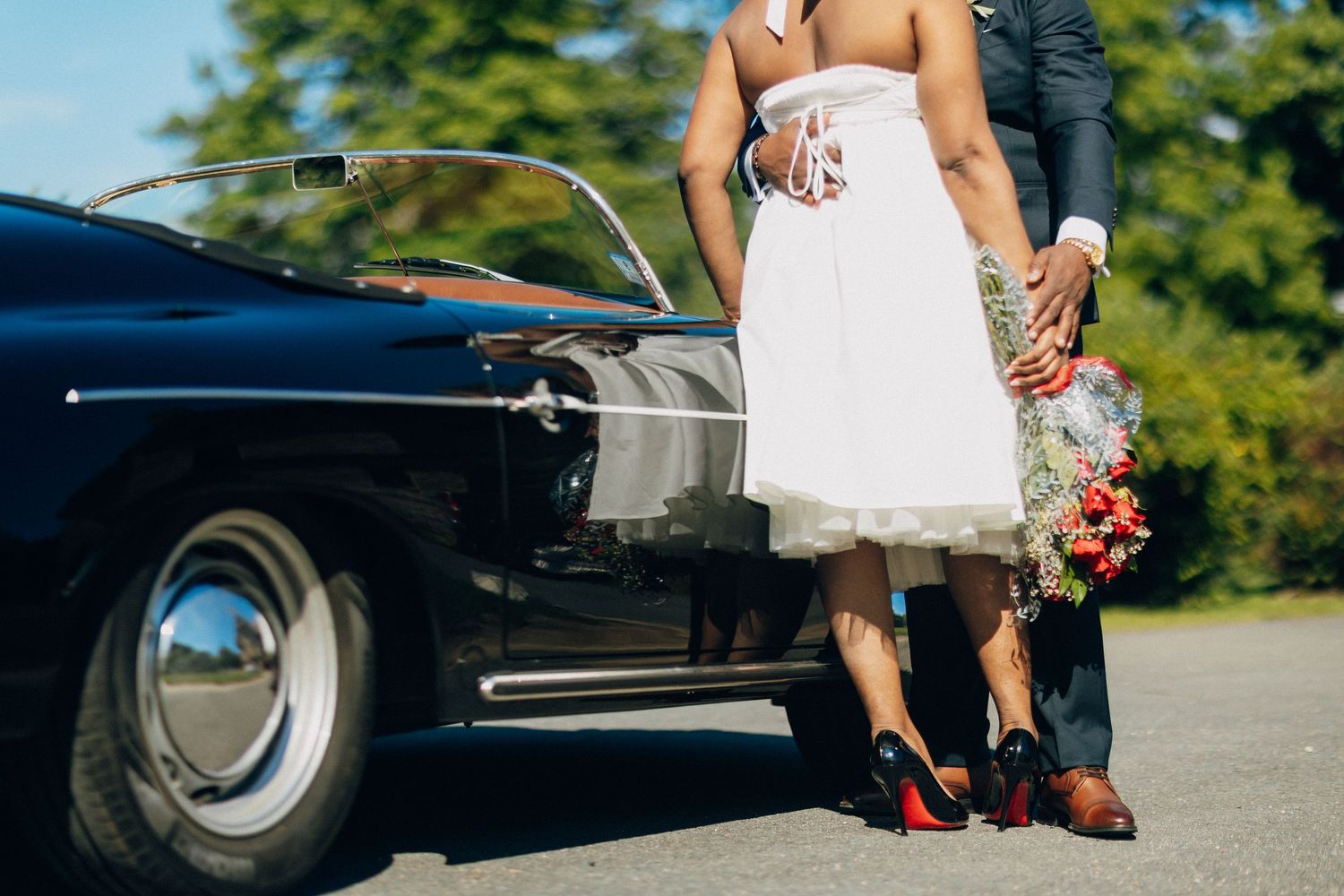 Close-up of bride in white dress and groom holding a bouquet beside a vintage black convertible.