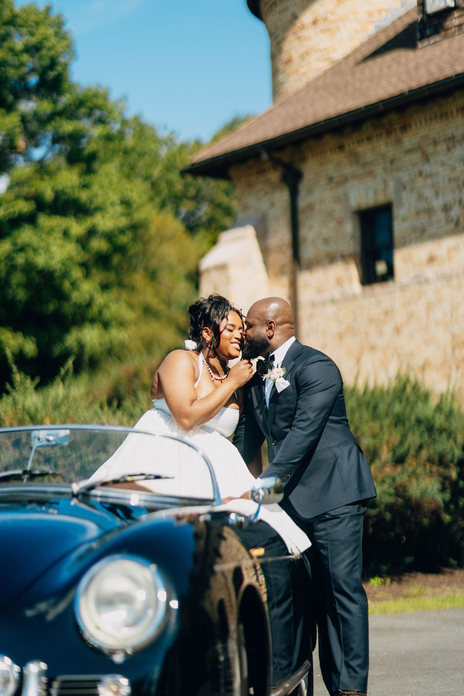 Bride sitting on hood of vintage black Porsche as groom leans in for a kiss near stone building.