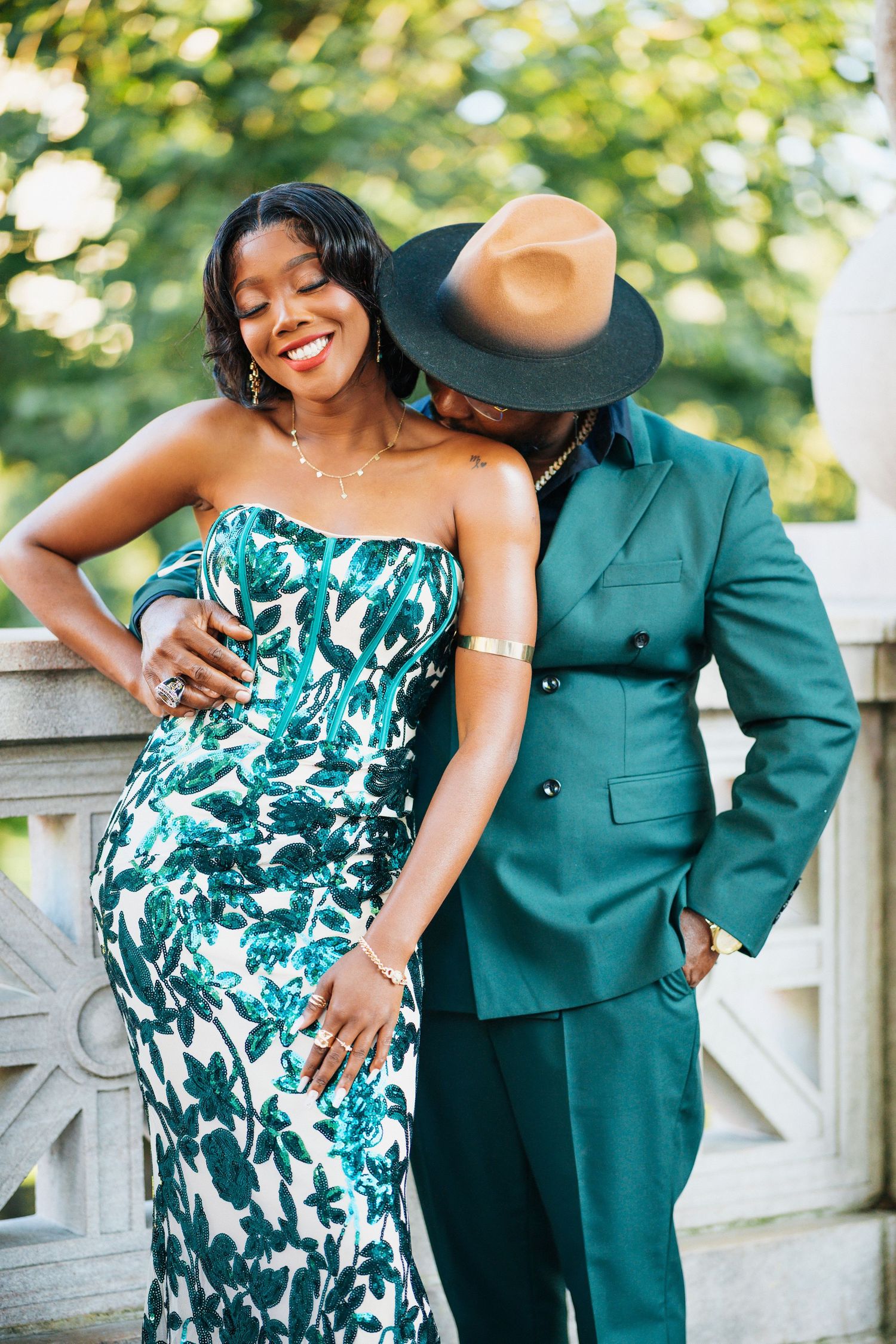 Elegant couple in matching green outfits posing outdoors, woman in floral gown and man in teal suit with wide-brim hat.